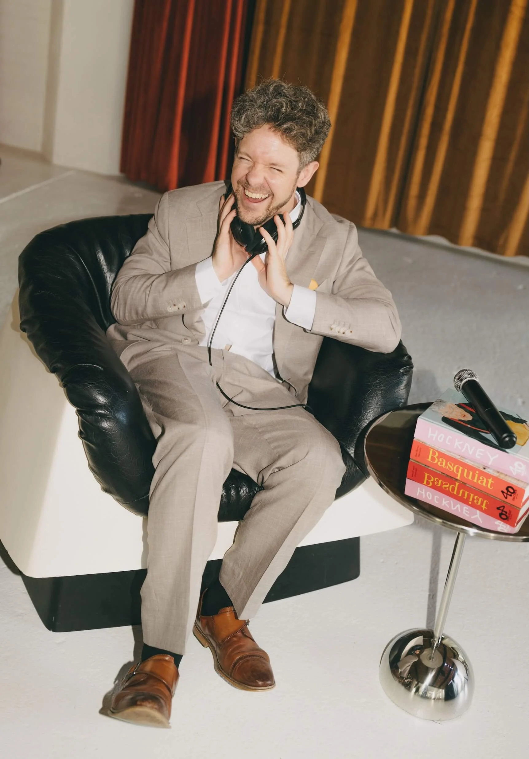 A man with curly hair and a beard, dressed in a beige suit and white shirt, sitting in a black leather chair, laughing and holding headphones to his ears. There are books stacked on a side table next to him and a microphone on a stand nearby.