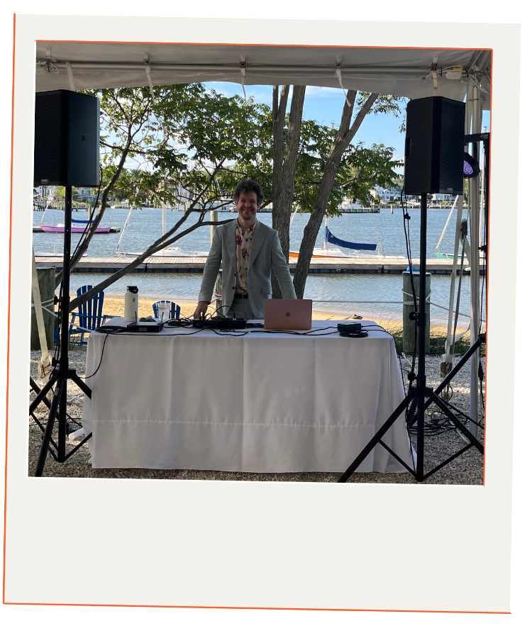 A DJ in a gray suit standing behind a DJ booth with audio equipment, set up under a canopy outdoors near a lake with sailboats and trees in the background.