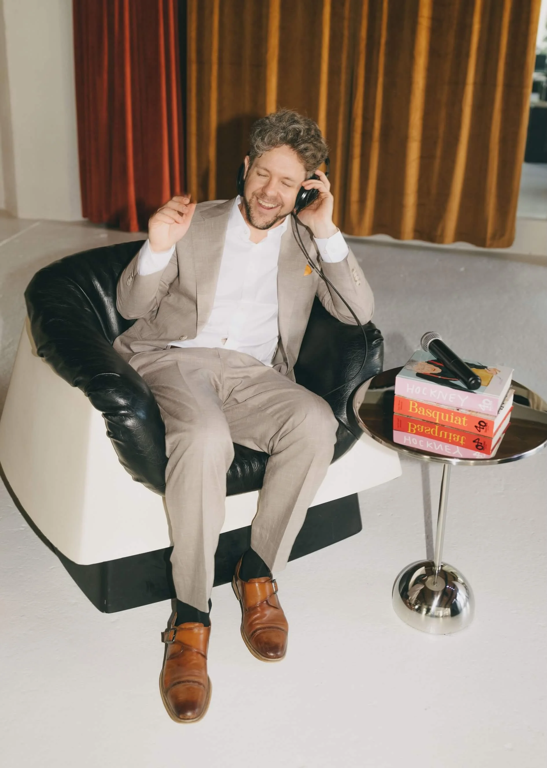 A man with curly hair and a beard, wearing a beige suit and white shirt, sitting in a black chair, smiling with eyes closed and holding headphones. Beside him is a small table with a few books and a microphone, with a background featuring brown wooden panels.
