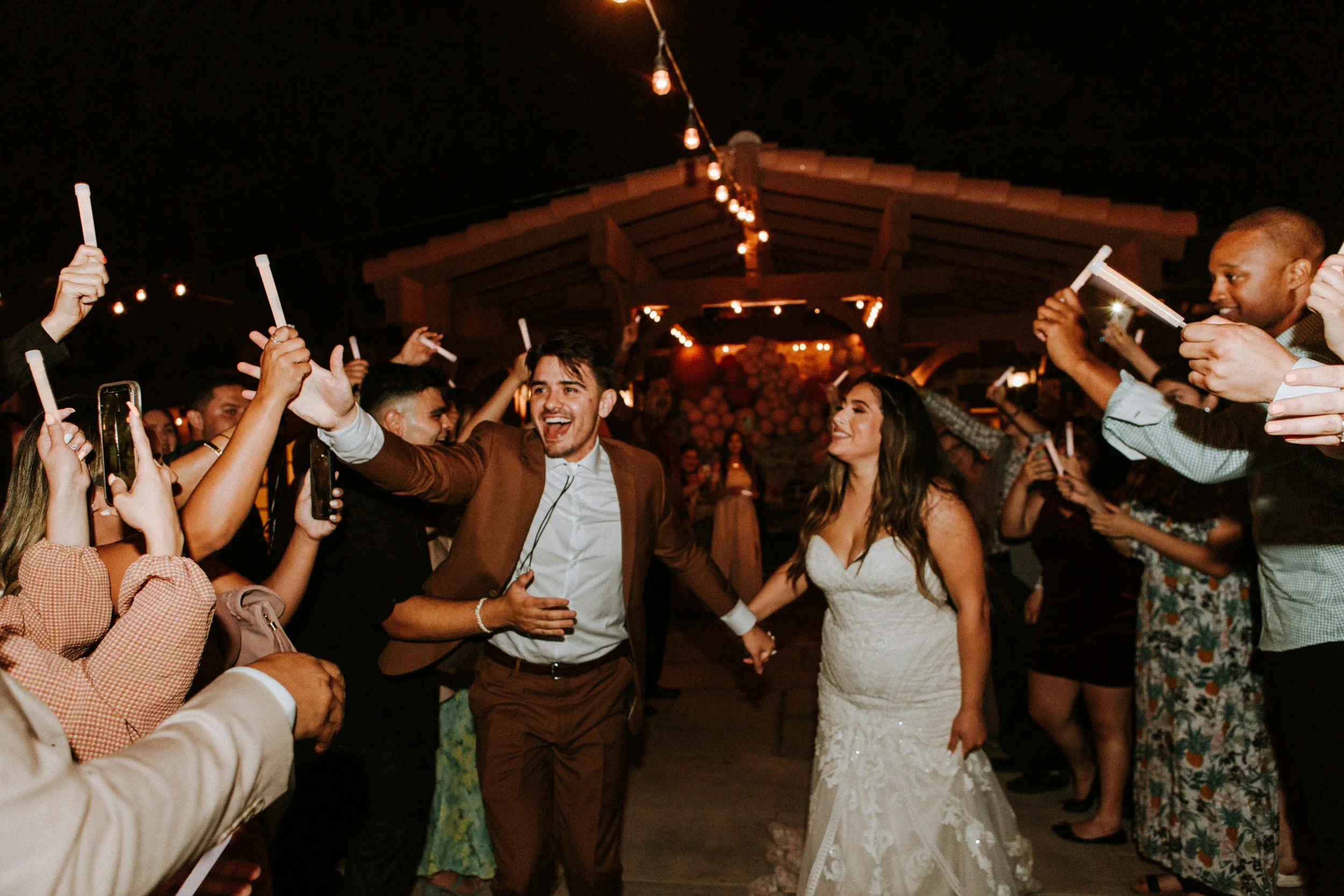 People dancing and enjoying themselves at an outdoor celebration at night, with string lights overhead and a wooden pavilion in the background, including a bride in a white dress.