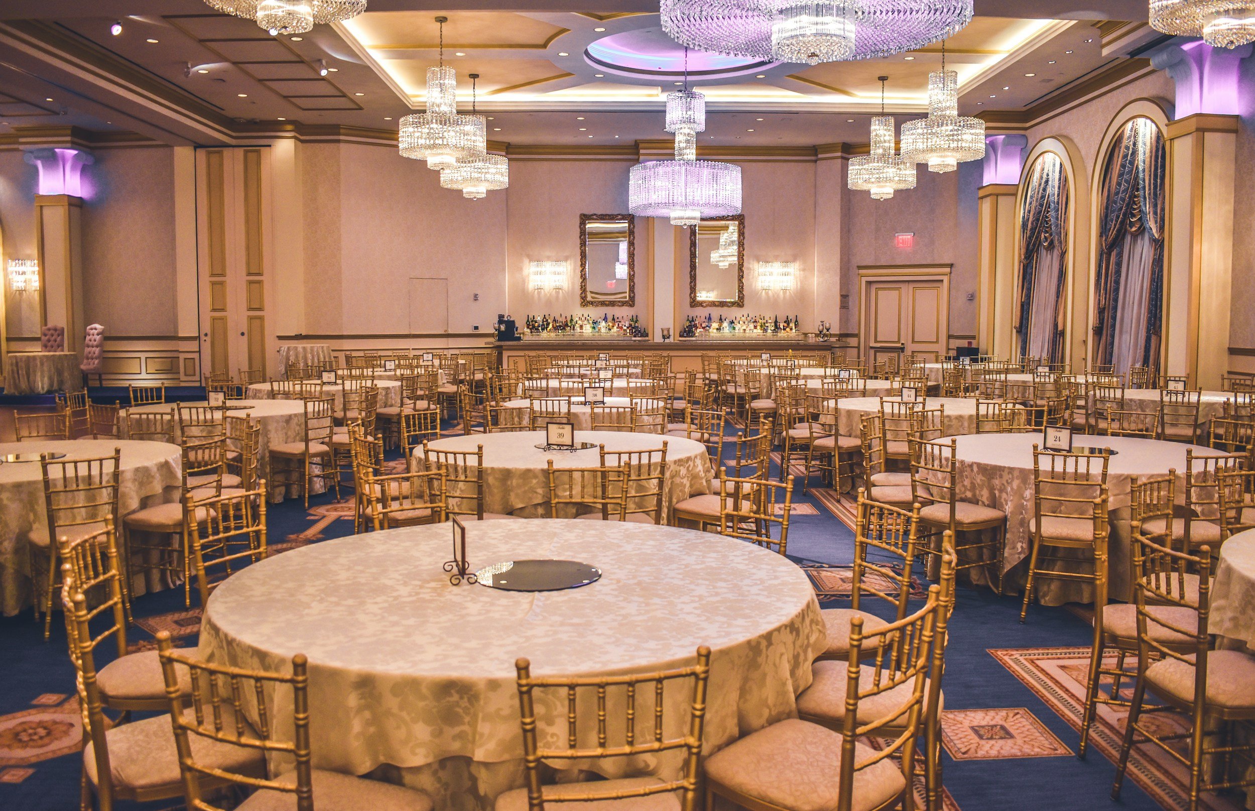 An elegant banquet hall decorated with multiple crystal chandeliers, gold Chiavari chairs around round tables draped with cream tablecloths, large mirrors on the back wall, and purple accent lighting on the walls.