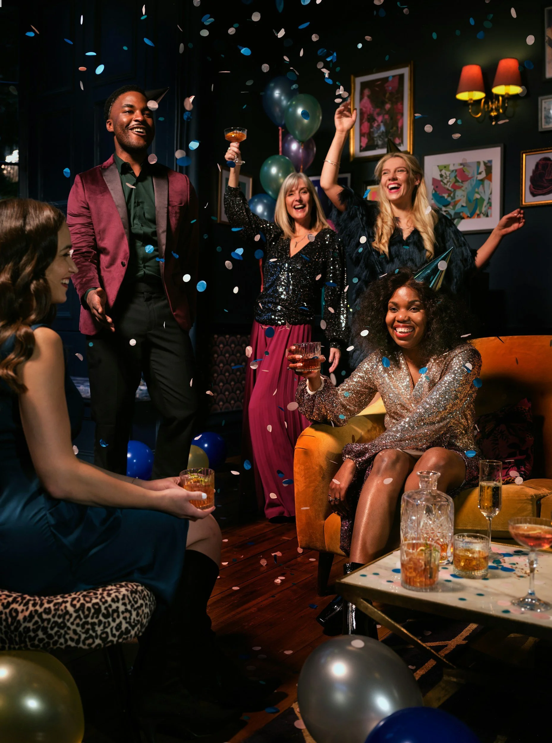 Group of five women and one man celebrating at a party with balloons and confetti, smiling and enjoying drinks in a decorated living room.