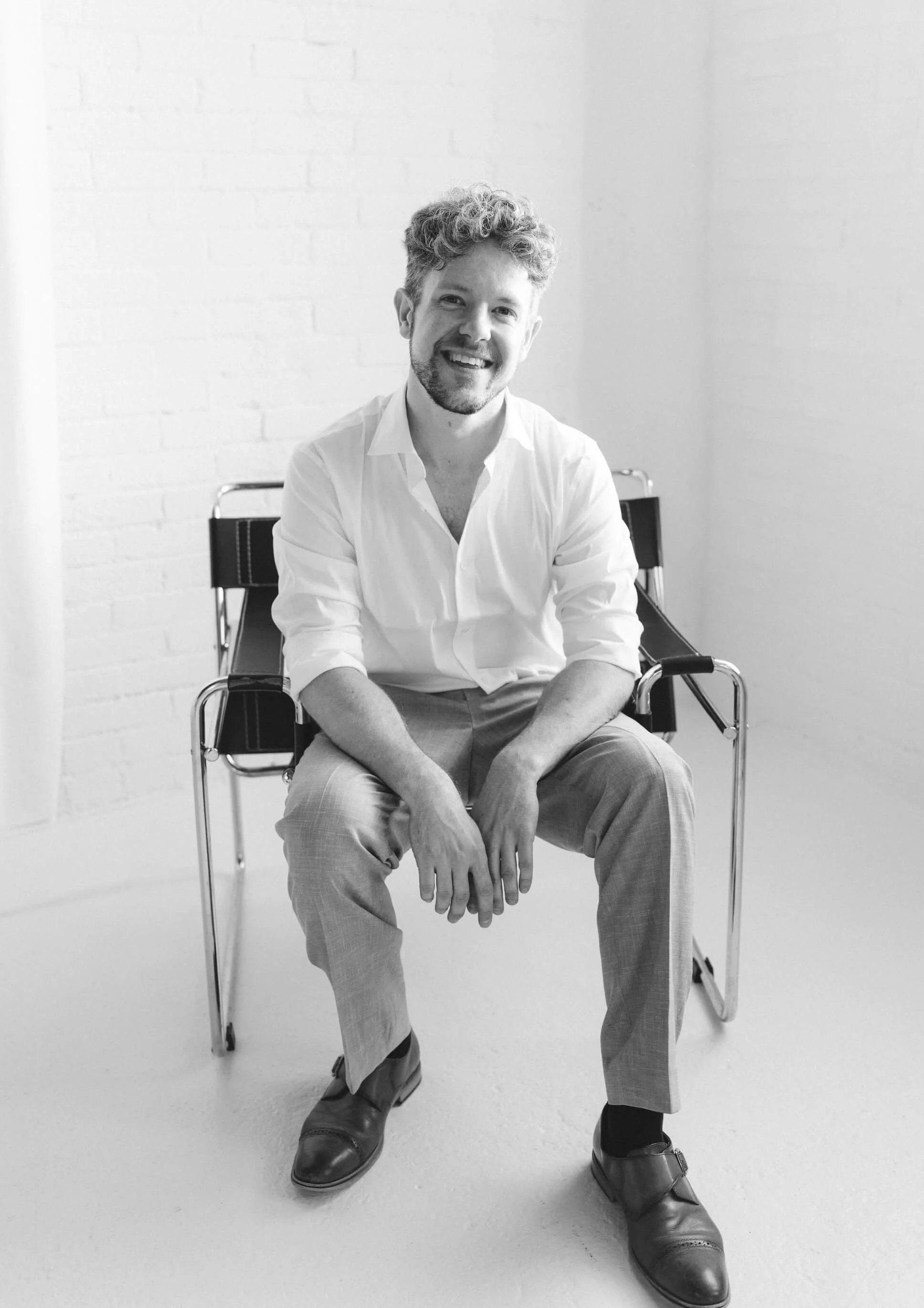 Black and white photo of a man sitting on a chair, smiling, wearing a white shirt, light pants, and dress shoes, in a room with white walls.
