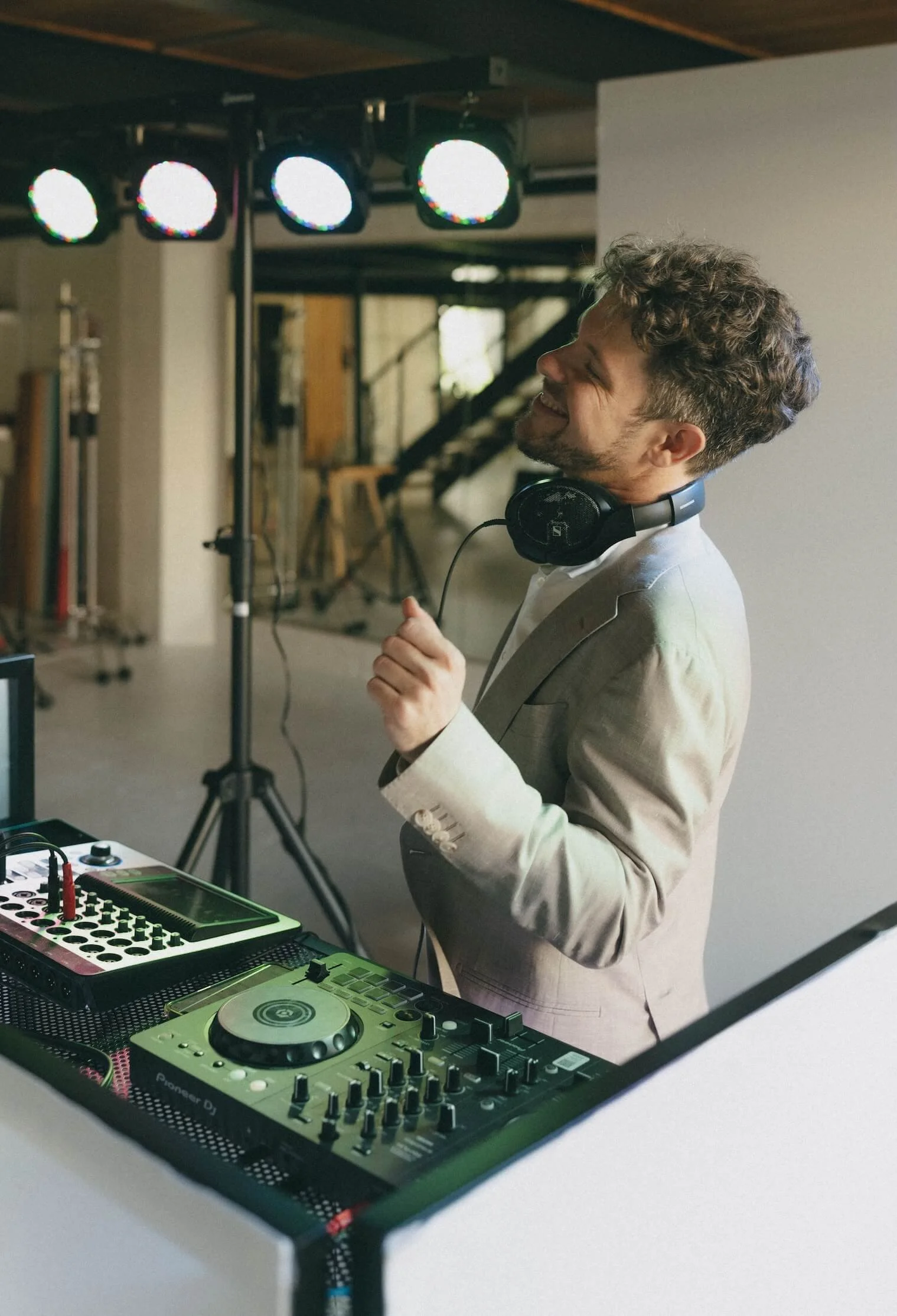 Man in a beige suit with headphones around his neck, smiling and dancing or DJing behind a DJ setup with turntables and audio equipment in a room with a wooden ceiling.