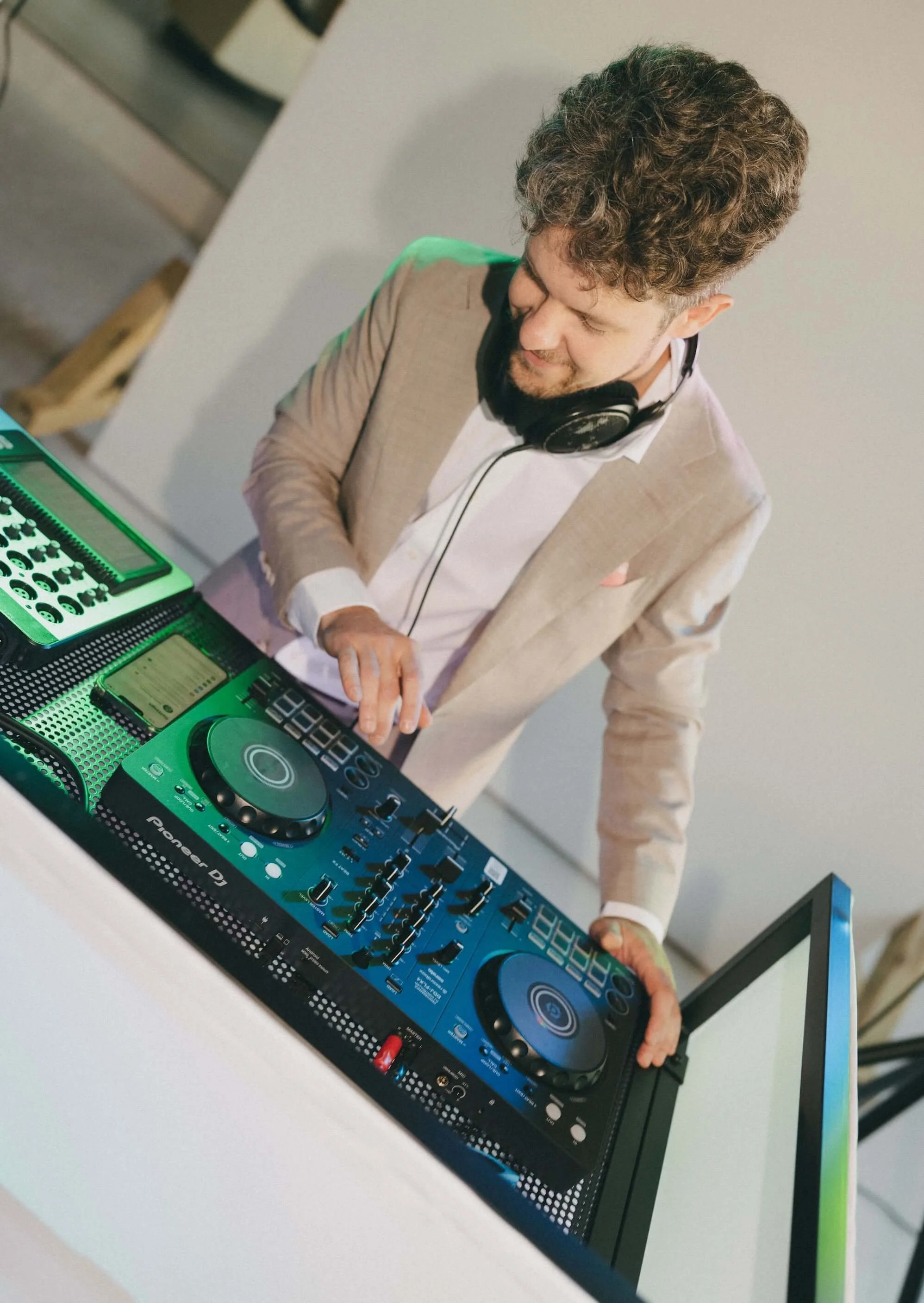 A man with curly hair wearing headphones and a beige blazer operating a DJ controller with green lighting effects.
