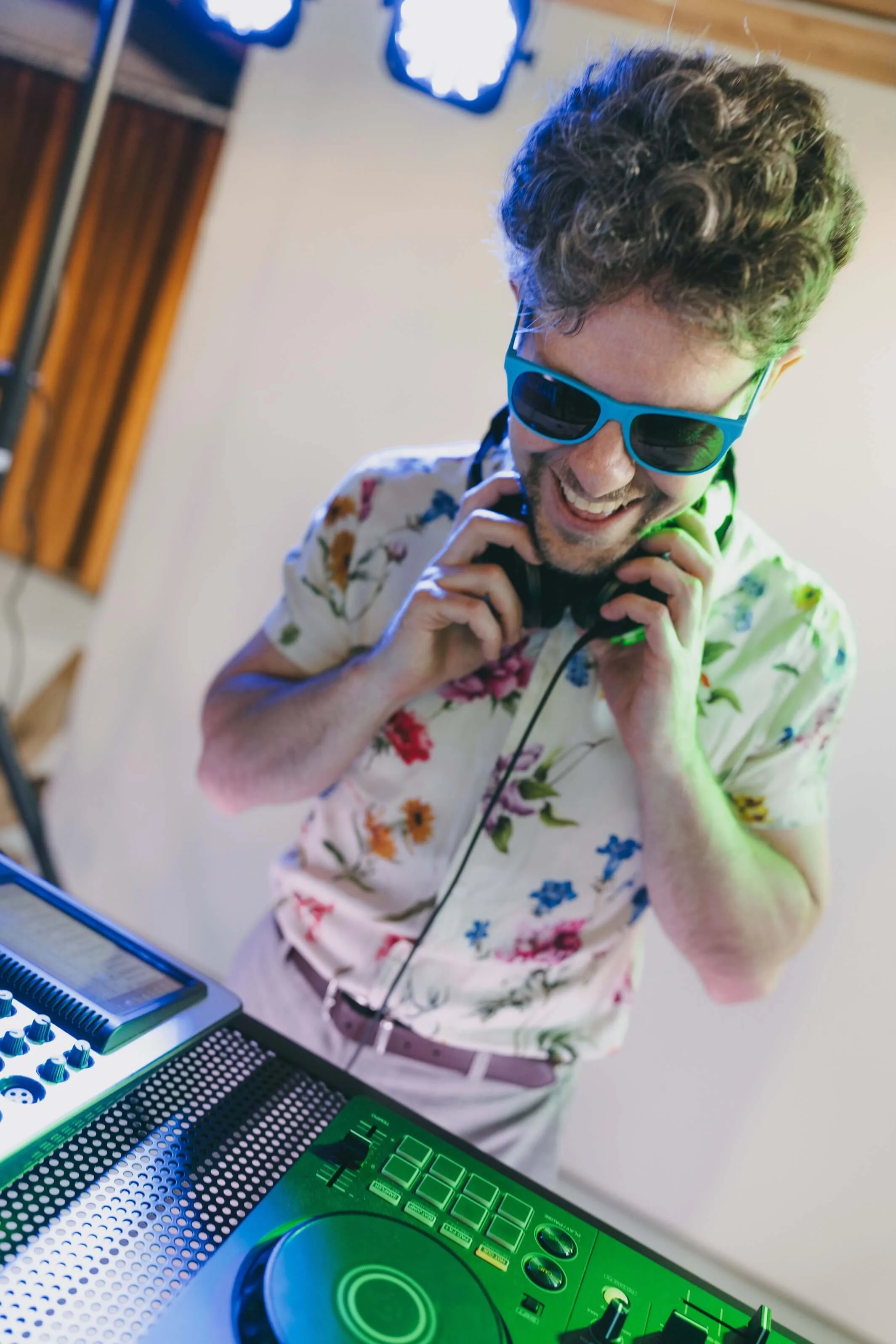 A man with curly hair wearing blue sunglasses and a floral shirt, smiling while holding headphones, in front of DJ equipment with a green digital display.