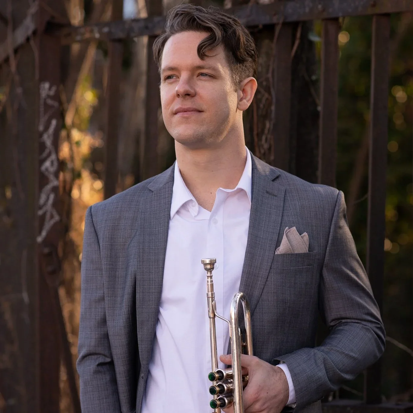 A man dressed in a gray suit and white shirt holding a trumpet, standing outdoors with a wooden fence and trees in the background.