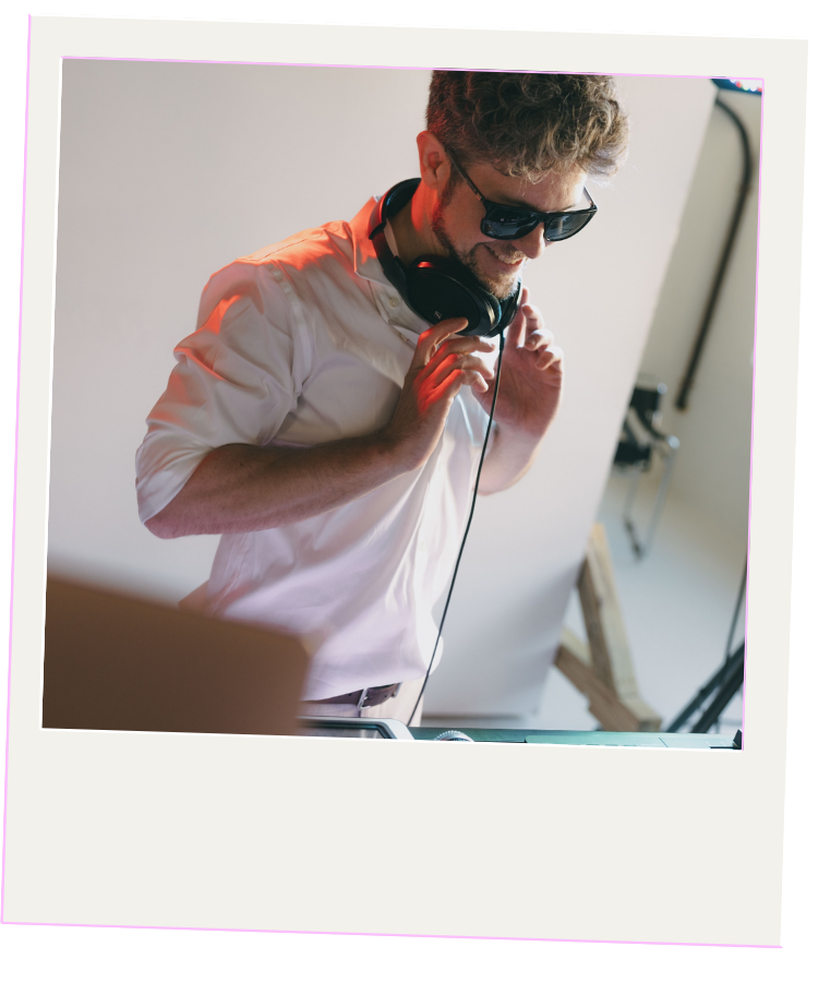 A young man wearing sunglasses and headphones around his neck, smiling and holding the headphone cord while standing in a room with a plain white wall.
