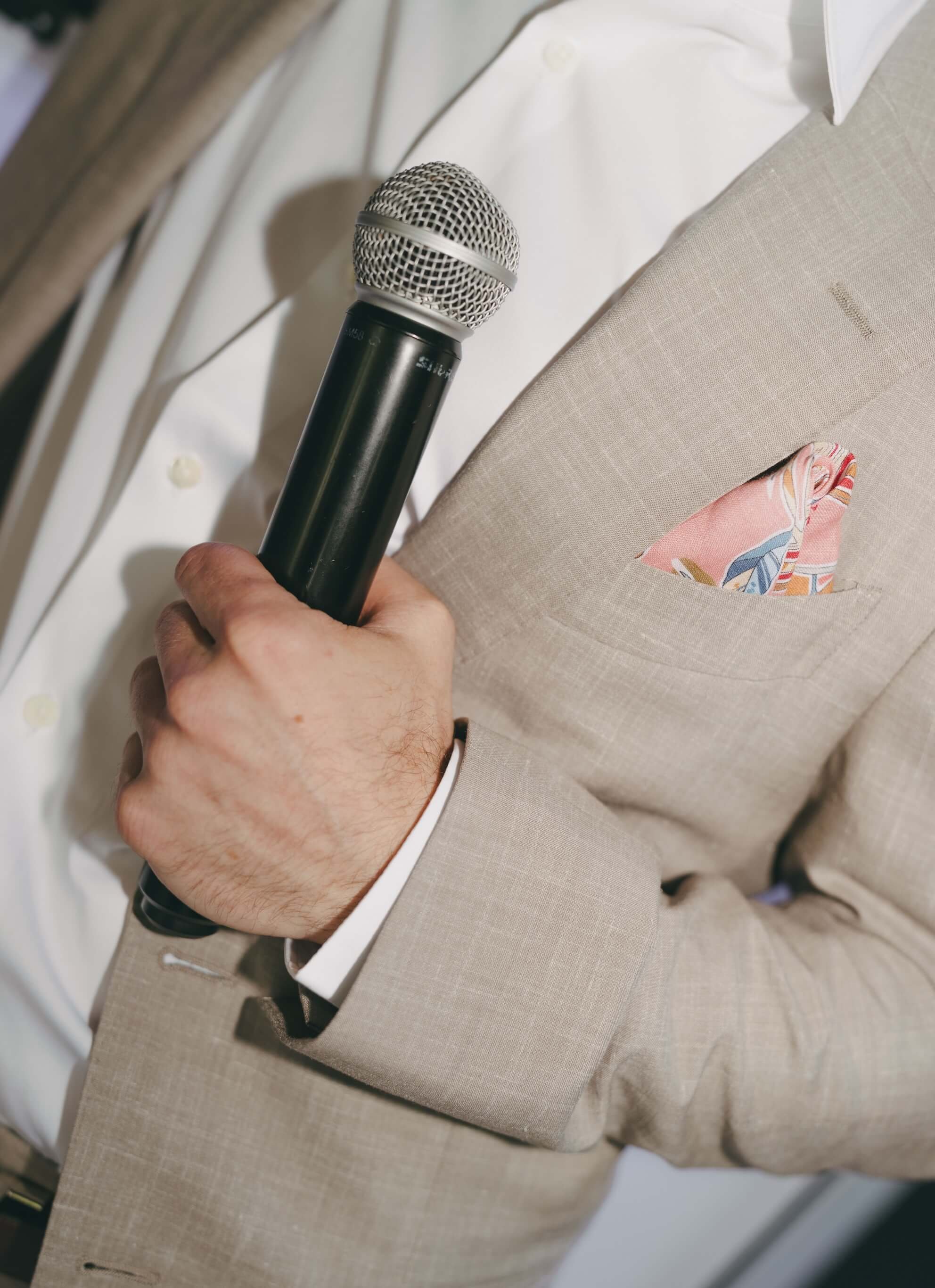 A man in a beige suit holding a microphone, with a beige pocket square with a colorful pattern in his breast pocket.