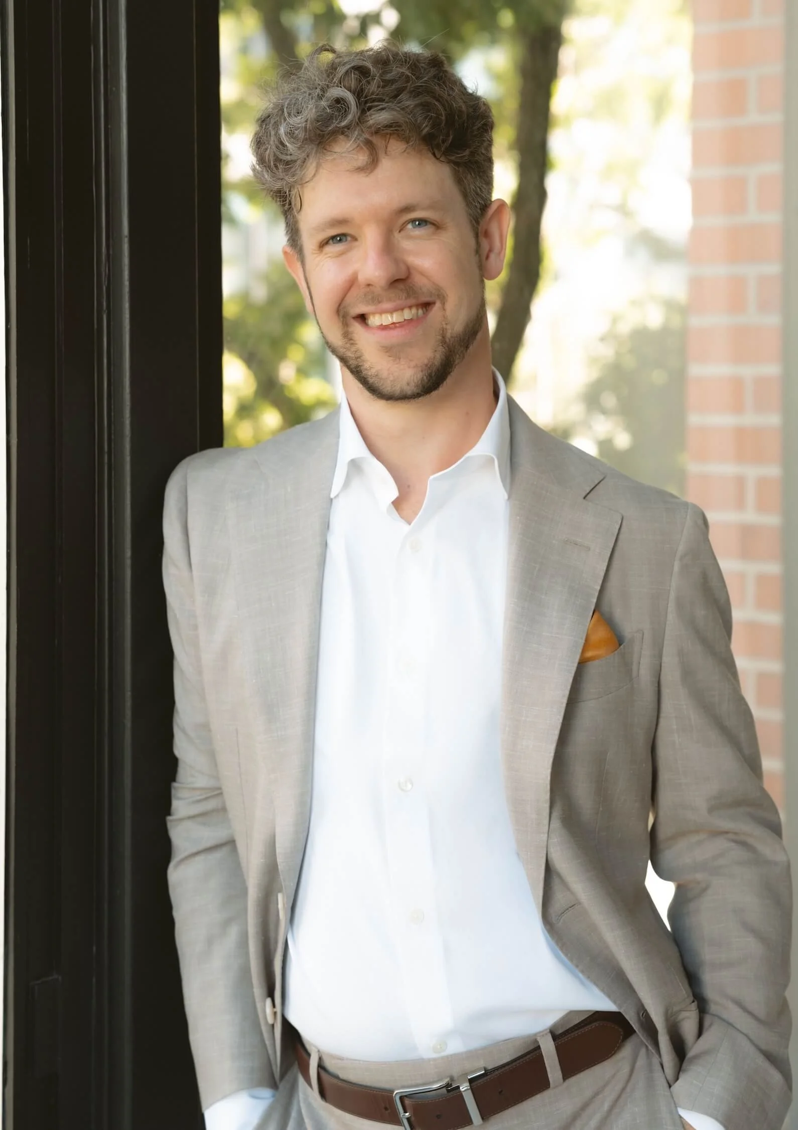 Young man with curly hair and a beard smiling, dressed in a light gray suit with a white dress shirt, standing outdoors near a brick building with trees in the background.
