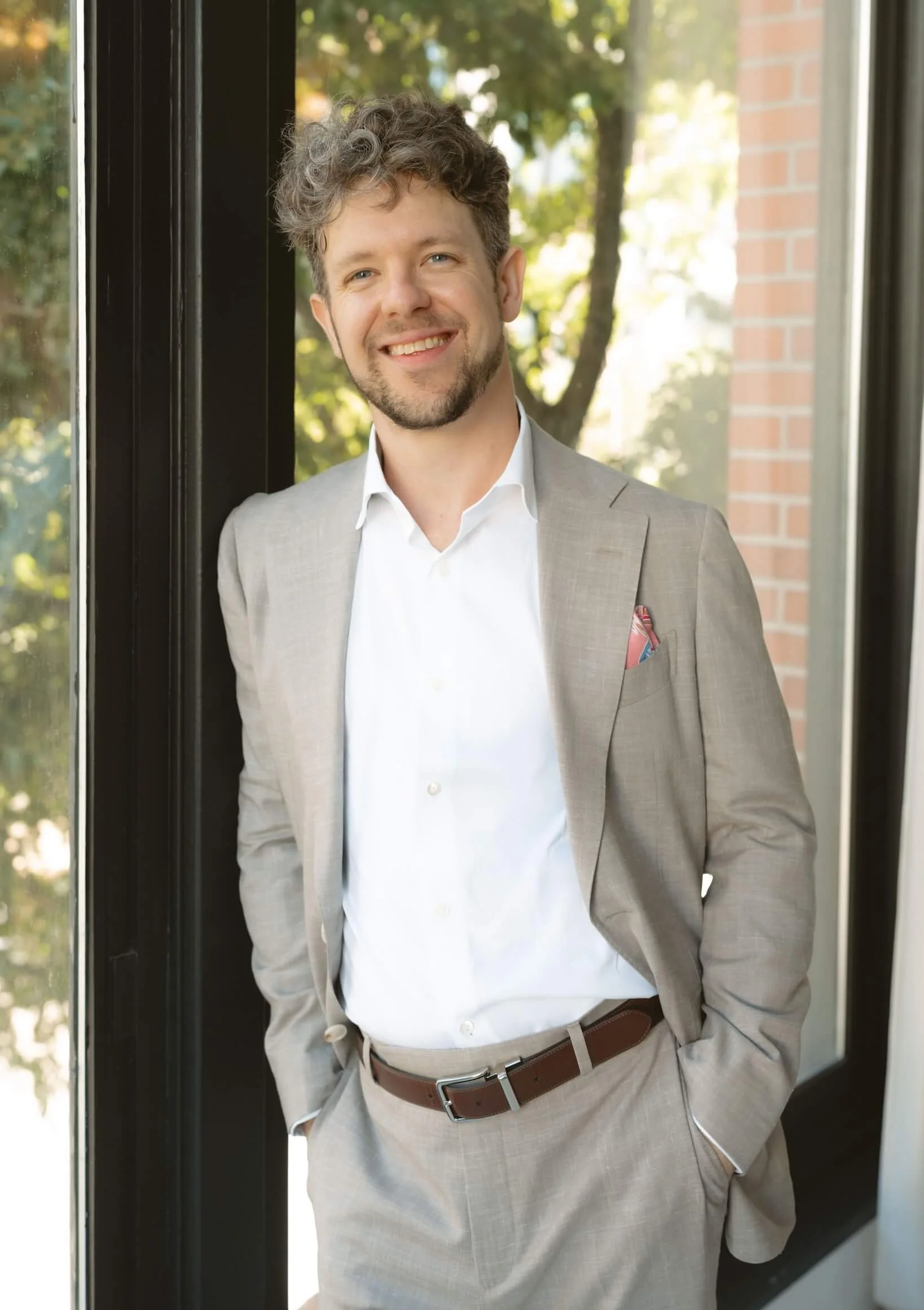 A man dressed in a light gray suit with a white shirt and a brown belt, standing outside a building, smiling, with trees and a brick wall in the background.