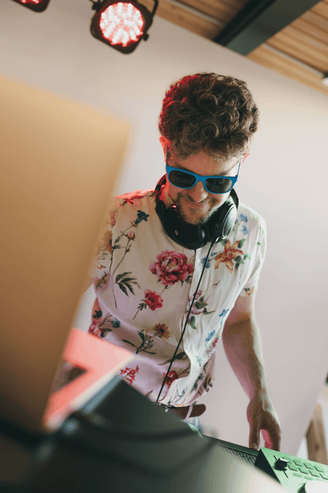 A man with curly hair wearing sunglasses and a floral shirt, smiling as he DJs at a mixing console, with headphones around his neck in a modern setting.