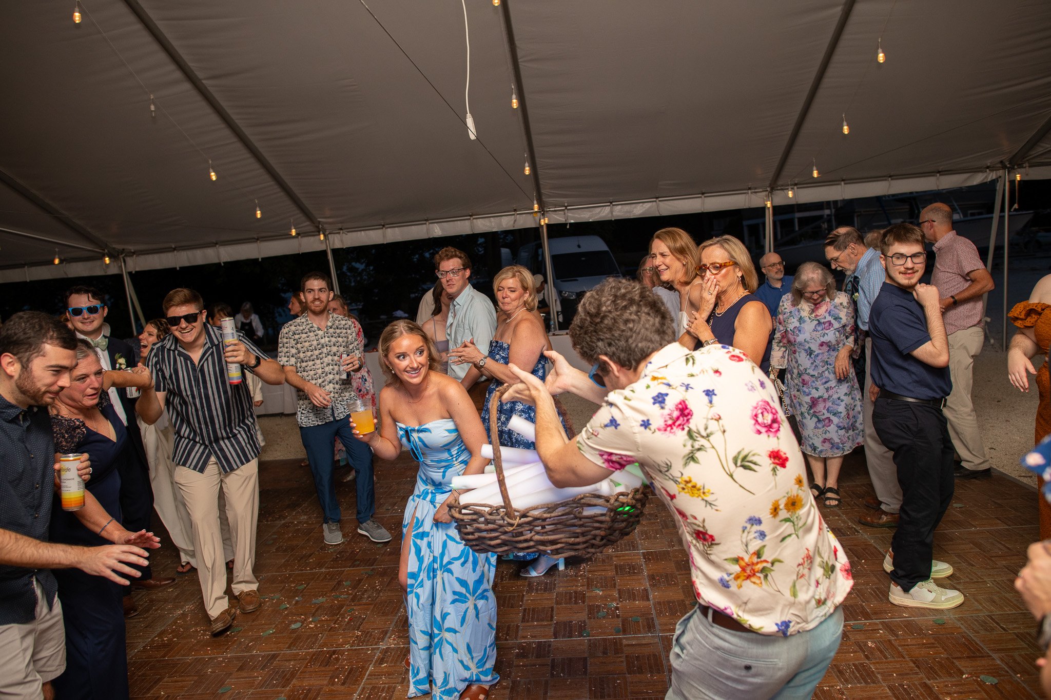 A group of people at a lively party or celebration under a large tent, dancing and laughing. The event features festive attire, drinks, and a cheerful atmosphere with string lights hanging from the ceiling.
