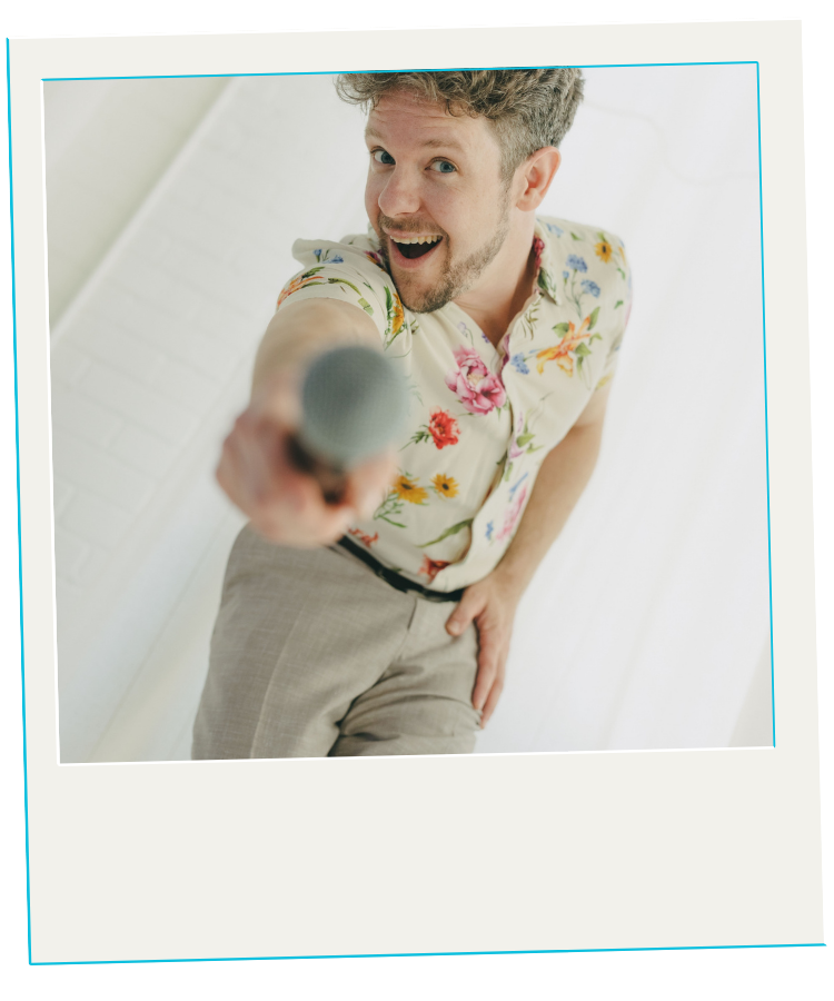 A man with curly hair and a beard smiling and holding a microphone towards the camera, wearing a floral shirt, standing against a bright background.