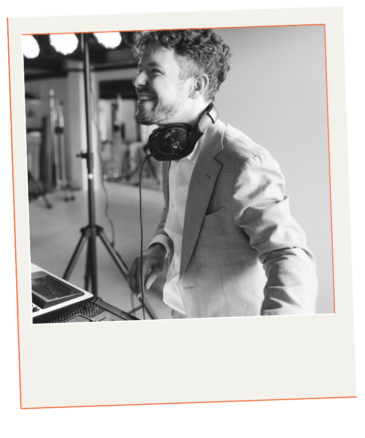A smiling man in a suit with headphones around his neck standing at a DJ booth in a studio or event space.