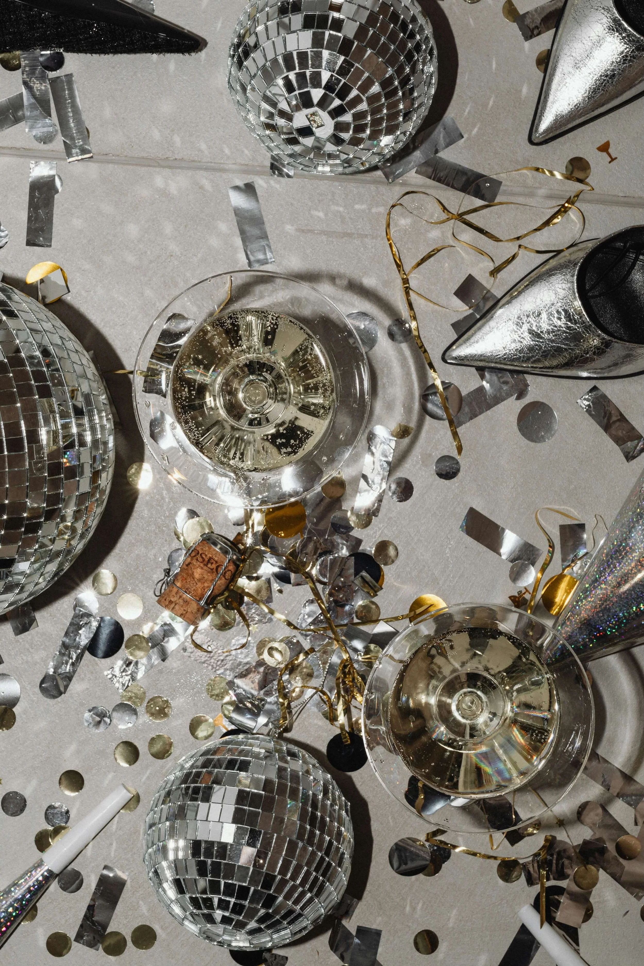 A scattered assortment of disco balls, champagne glasses, metallic ribbons, and confetti on a light-colored surface, suggesting a celebration or party setup.