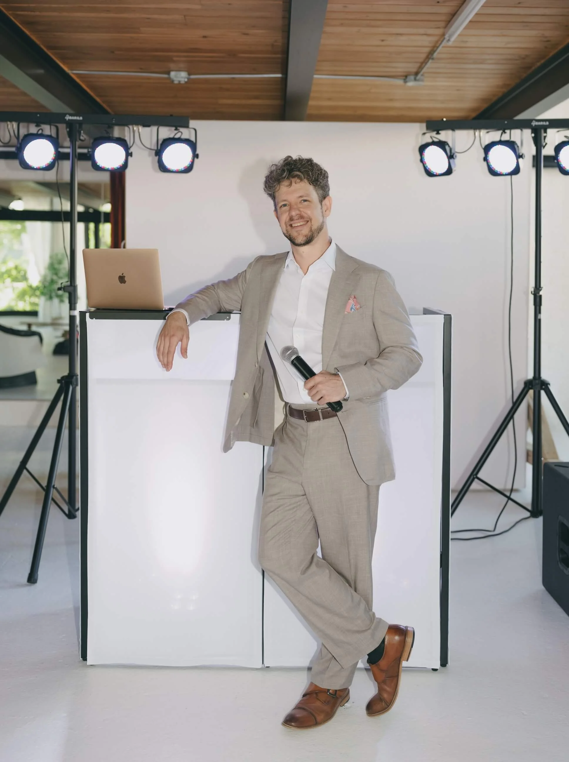 A man in a light gray suit with a white shirt, holding a microphone, smiling, and posing with one foot on a white box. Behind him are stage lights, a wooden ceiling, and large windows letting in natural light.