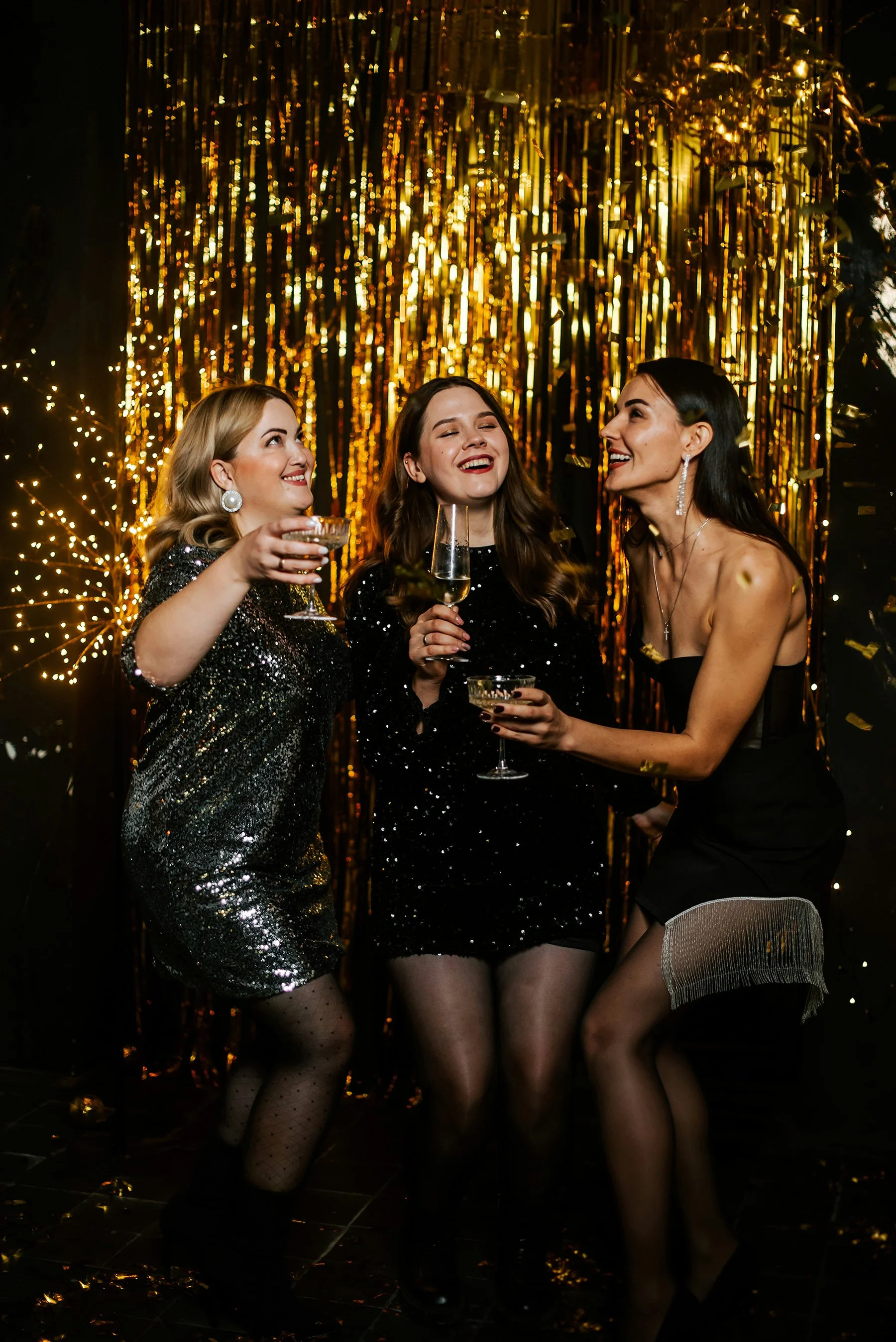 Three women dressed in glamorous black and gold party outfits celebrating at a festive event with gold curtain backdrop and confetti on the floor. They are holding champagne glasses and smiling.
