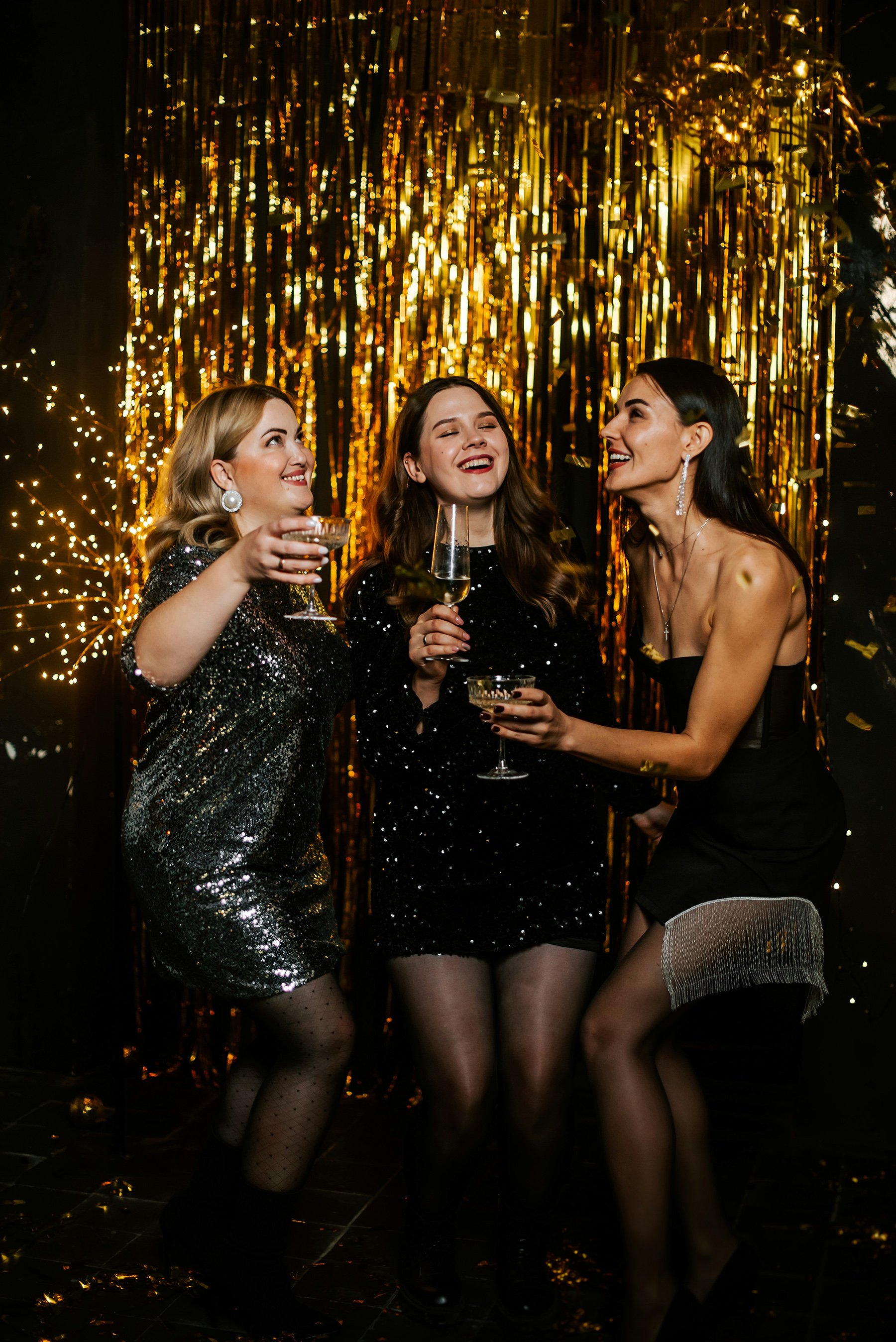Three women dressed in party attire are celebrating together with drinks in hand, standing in front of a shiny gold tinsel backdrop at a party or event.