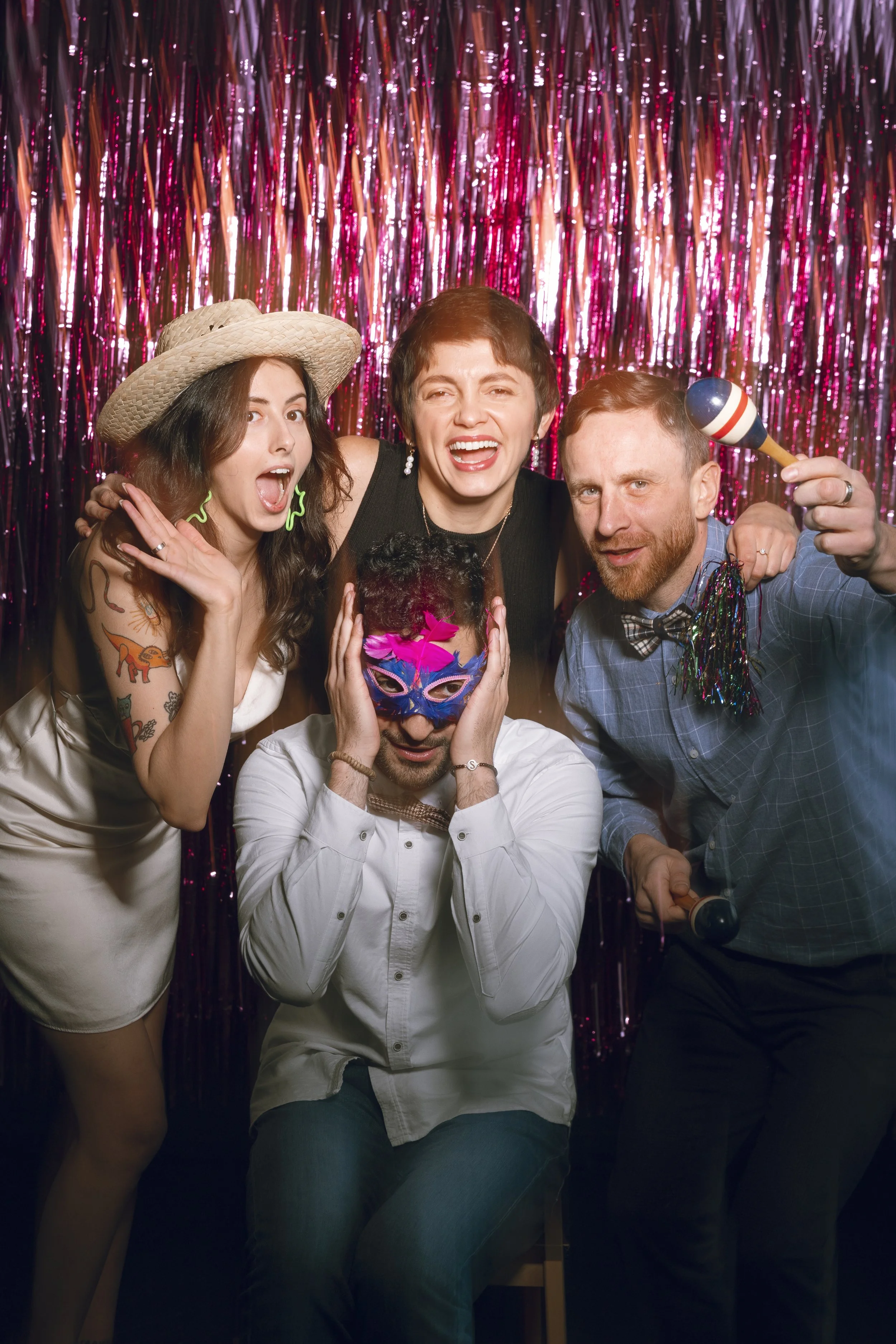 Group of five friends at a party, wearing fun accessories like a sunhat, mask, and maracas, in front of pink metallic streamers.