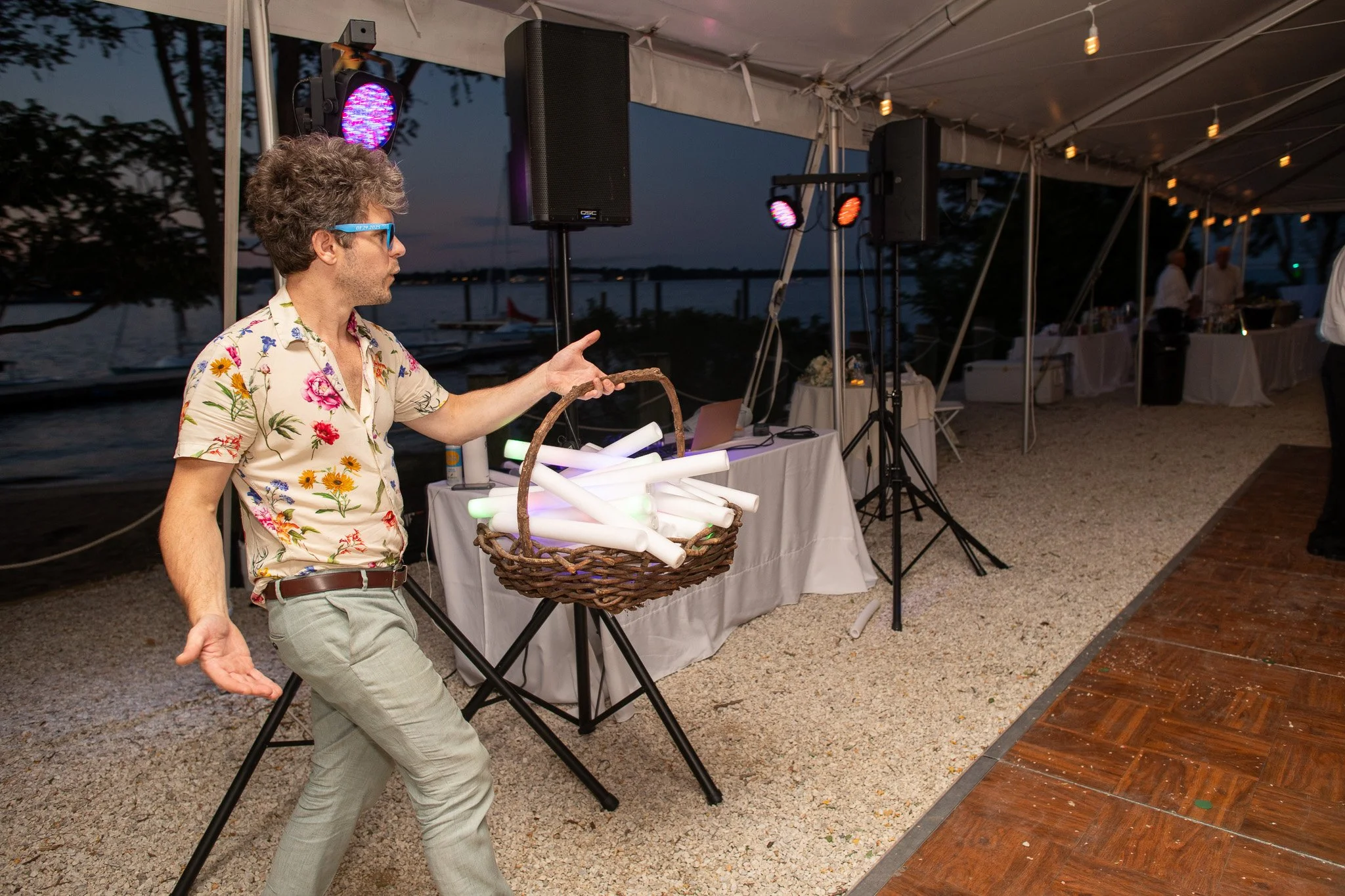 A man with curly hair, wearing glasses, a floral shirt, and light-colored pants, is at an outdoor evening event under a tent. He is holding a basket filled with glowing objects and appears to be in motion, possibly dancing or gesturing. The background includes a table with items, speakers, and colorful lighting, with a body of water and boats visible beyond.