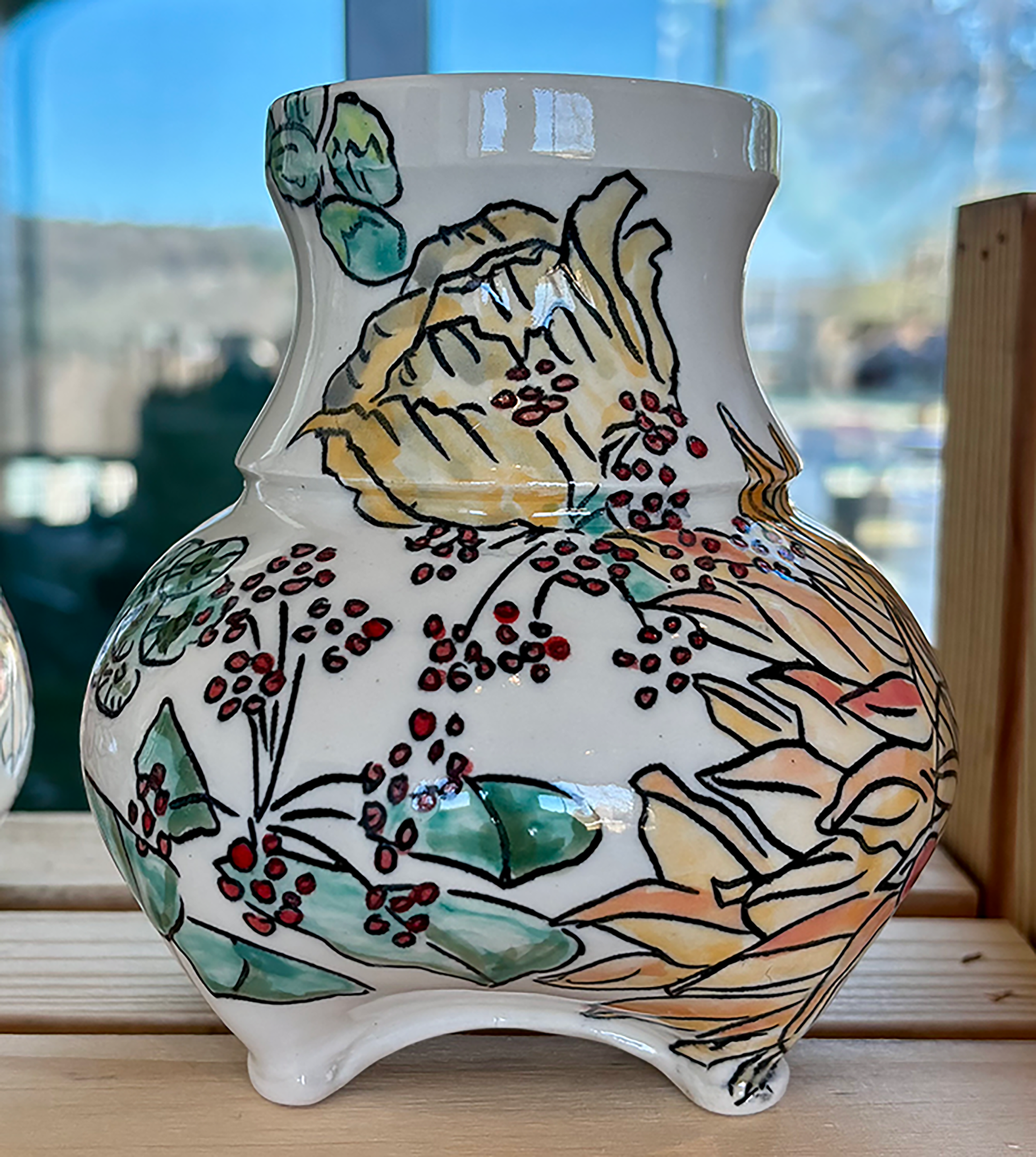 Decorative ceramic vase with a painted floral design, placed on a wooden surface with a bright window in the background.