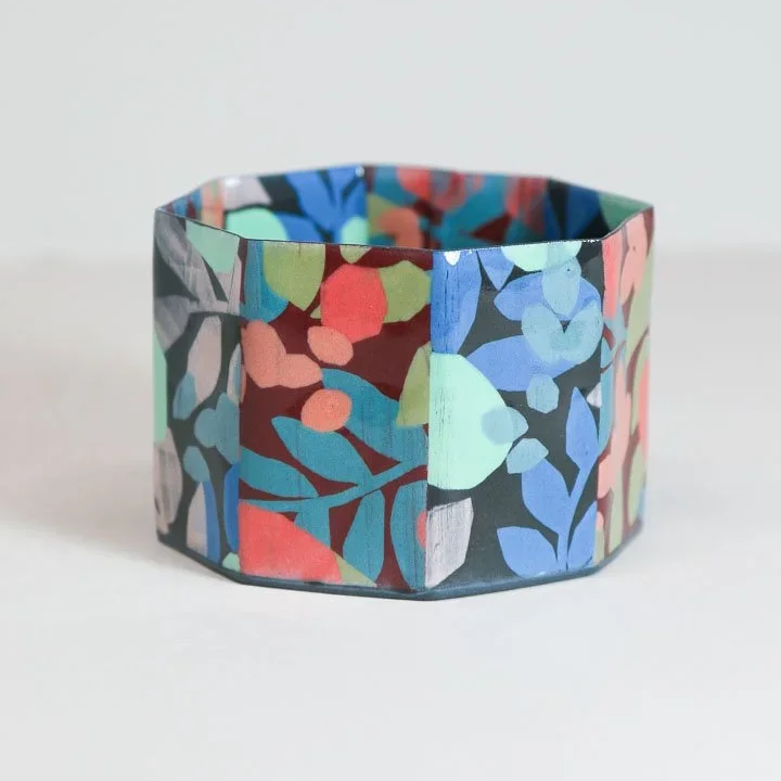 A colorful, patterned paper bracelet with a floral or abstract design in various shades of blue, green, pink, red, and gray, resting on a white surface by Lydia Johnson