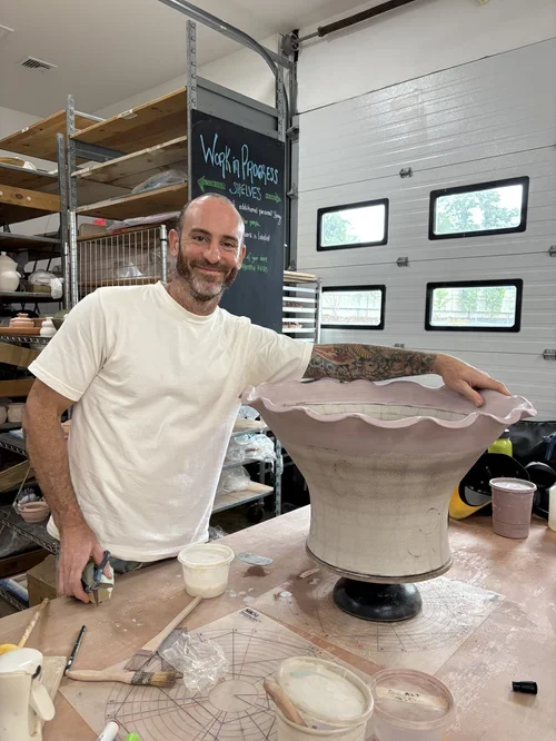 Pedro Centeno in a ceramics studio, posing next to a large, partially finished ceramic piece. The studio has shelves with pottery and tools, with a chalkboard in the background that says 'Work in Progress'.