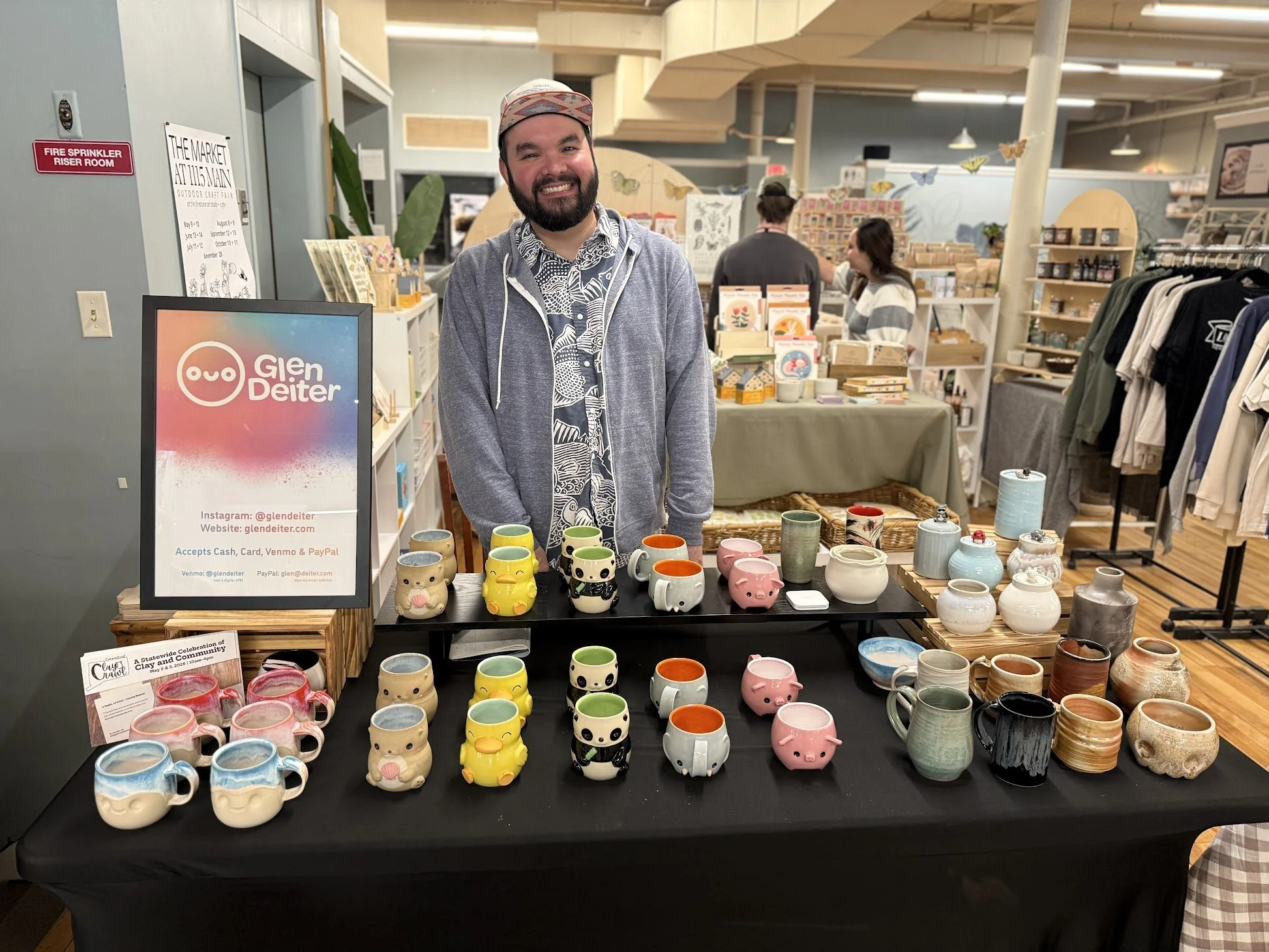 Potter and instructor at Silk City Clay in Manchester CT, Glen Deiter a man with a beard and a colorful cap smiling at a booth with colorful ceramic mugs shaped like animals and faces, at an outdoor market or craft fair.