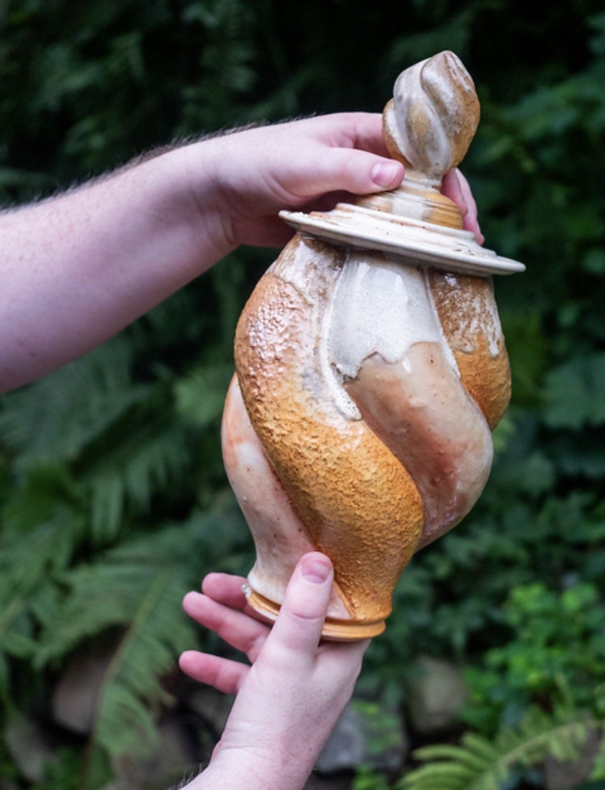 Hand holding a large, uniquely shaped ceramic jar outdoors by Kevin Shaw with green foliage in the background.