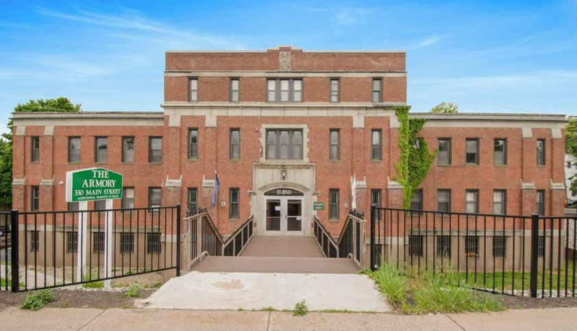 Brick building with green sign that reads "The Armory, 330 Main Street." Entrance ramp with black metal railing leading to the front door, green landscaping, and a clear blue sky.