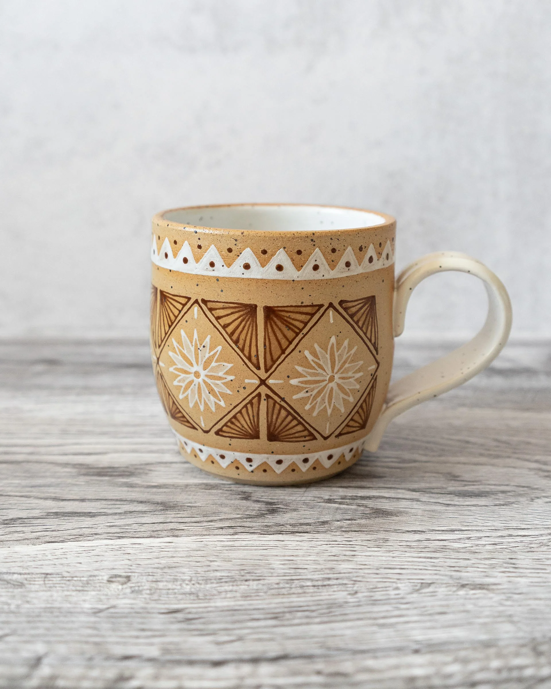 Close-up of a ceramic mug by Amy Brockman with geometric and floral patterns, placed on a wooden surface with a plain light-colored background.