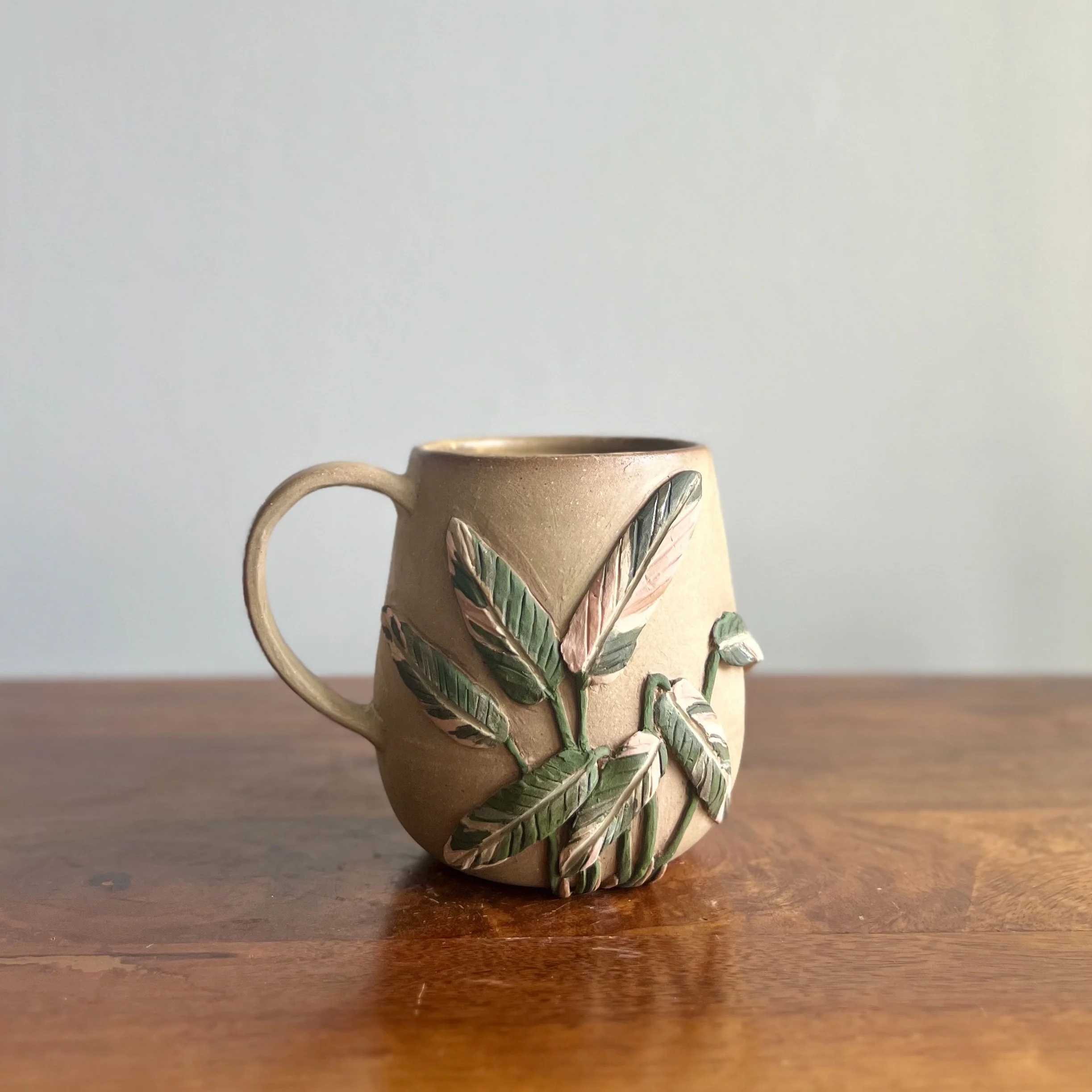 A ceramic mug with a handle, decorated with painted green and pink leaves and vines, resting on a wooden surface by Gleidymar Rivera