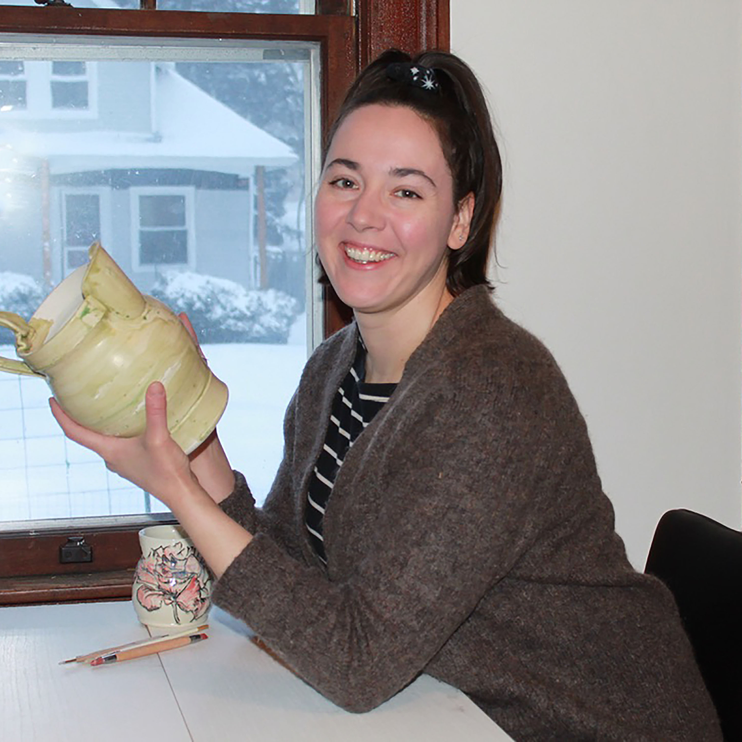 A woman with short dark hair, wearing a striped shirt and brown sweater, smiling and holding a ceramic mug, sitting at a table by a window with snow outside.