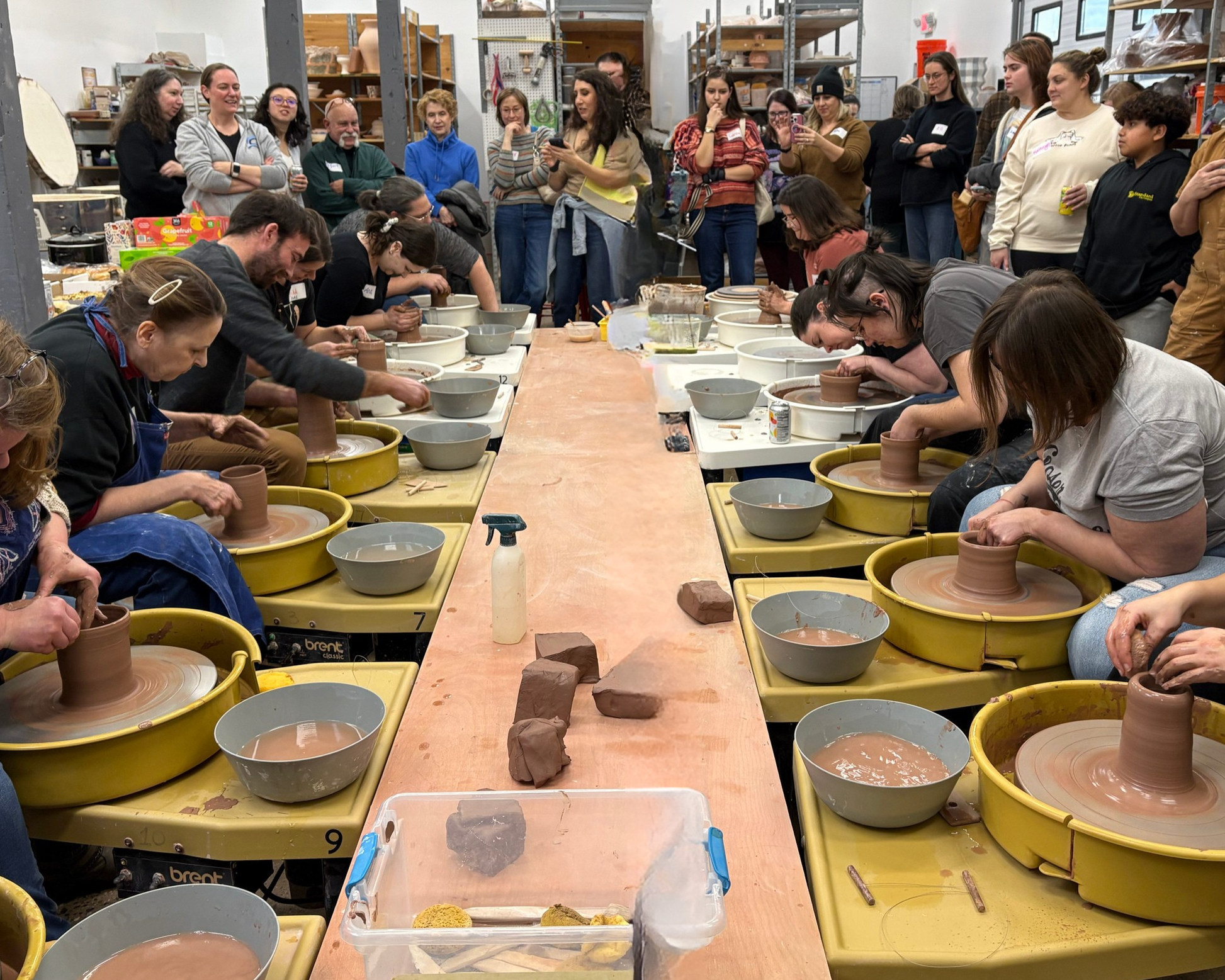 People are gathered around multiple pottery wheels in a ceramics class, shaping clay into pottery, with a crowded workshop filled with shelves of supplies and a group of onlookers observing.