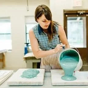 Lydia Johnson, A young woman pouring teal-colored paint from a container onto a surface in an art studio.