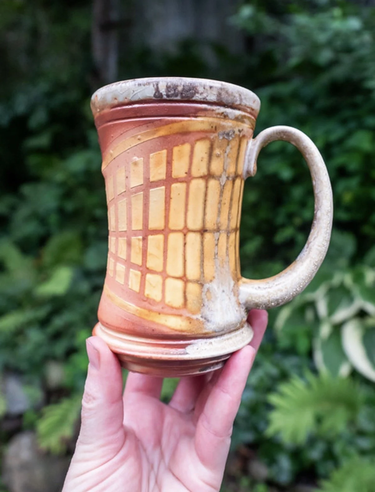 A hand holding a ceramic mug by Kevin Shaw with a geometric pattern, outdoors with green foliage in the background.