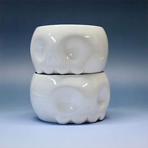 A pair of white ceramic skull-shaped objects stacked on top of each other against a blue gradient background. - Glen Deiter