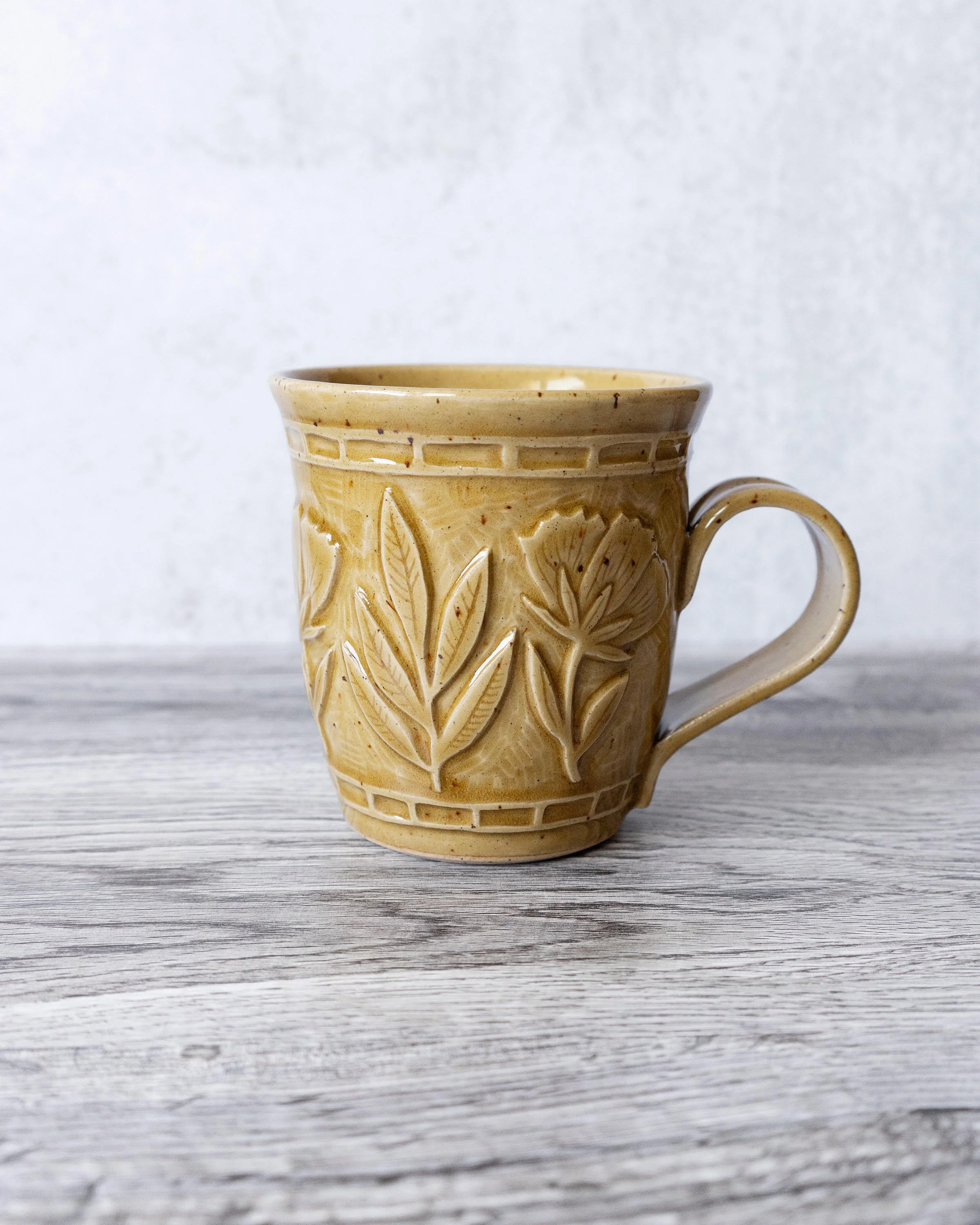 A beige ceramic mug by Amy Brockman with a leaf pattern design on the exterior, placed on a light-colored wooden surface with a plain white background.