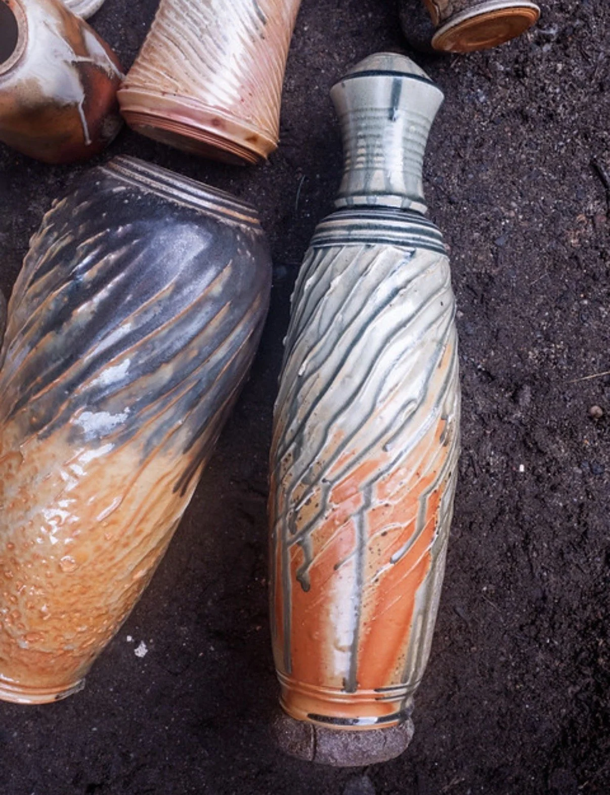 Close-up of several colorful, textured ceramic vases by Kevin Shaw with orange, black, white, and beige glaze designs on a dirt ground.