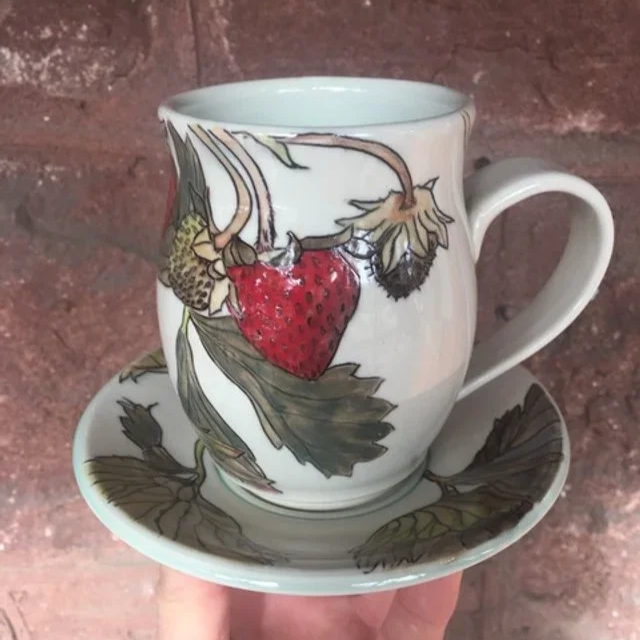 A ceramic mug with a strawberry design, placed on a matching saucer with leaf patterns by Pedro Centeno