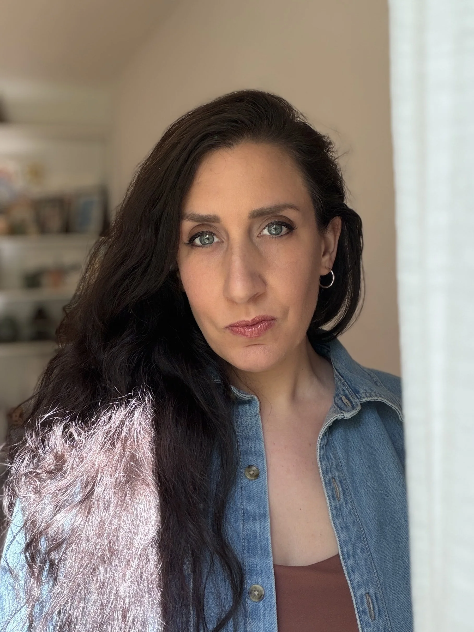 Stephanie Haines, owner of Silk City Clay in Manchester CT and founder of The Connecticut Clay Guild, a woman with dark hair and blue eyes, wearing a denim jacket, standing near a window with sheer curtains, with a blurry bookshelf in the background.