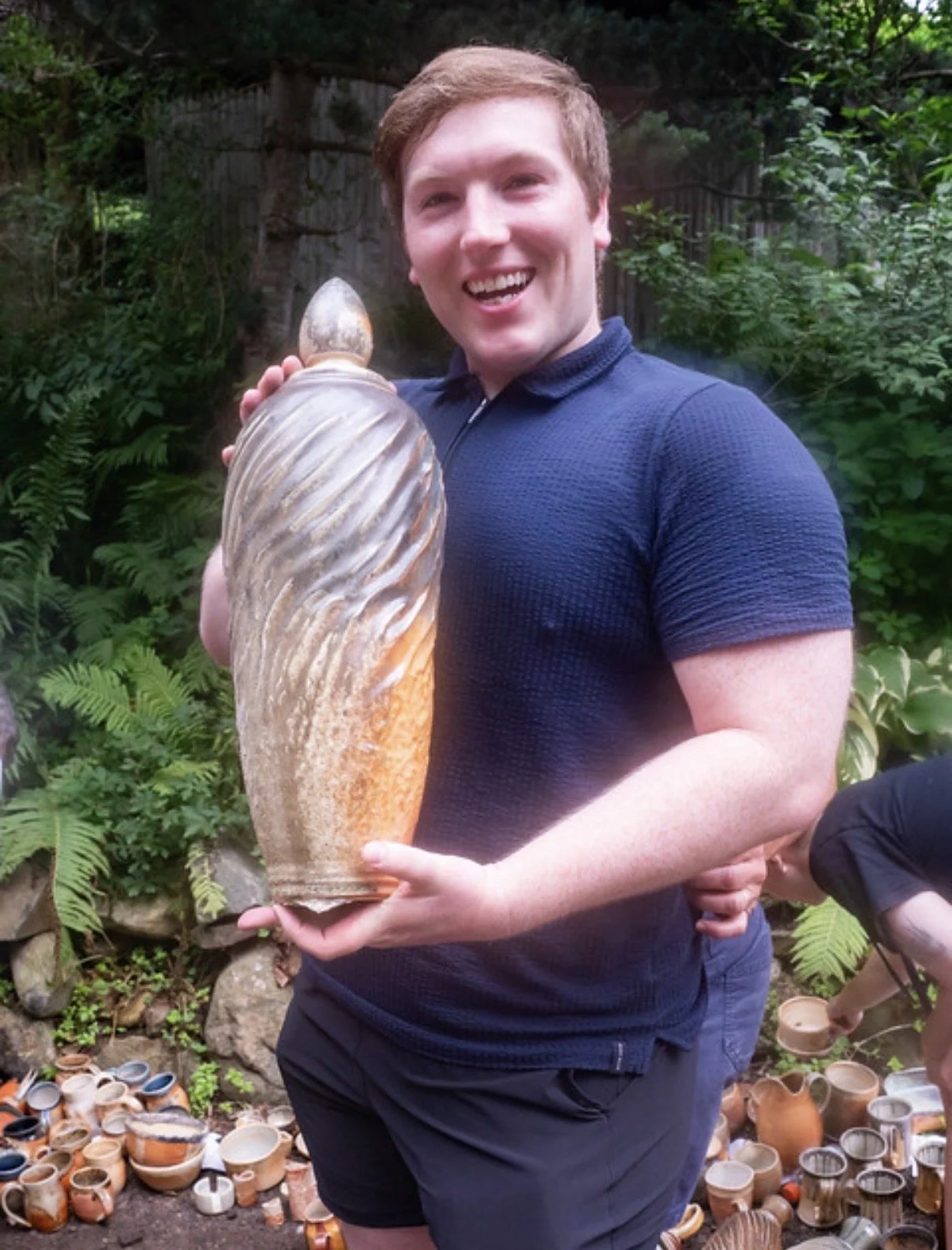 Connecticut potter and instructor at Silk City Clay in Manchester CT, Kevin Shaw. A young man smiling and holding a large conch shell outdoors surrounded by plants and pottery.