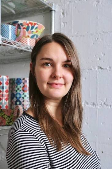 Lydia Johnson, A woman with long brown hair wearing a black and white striped shirt, standing indoors in front of shelves with colorful bowls and items.