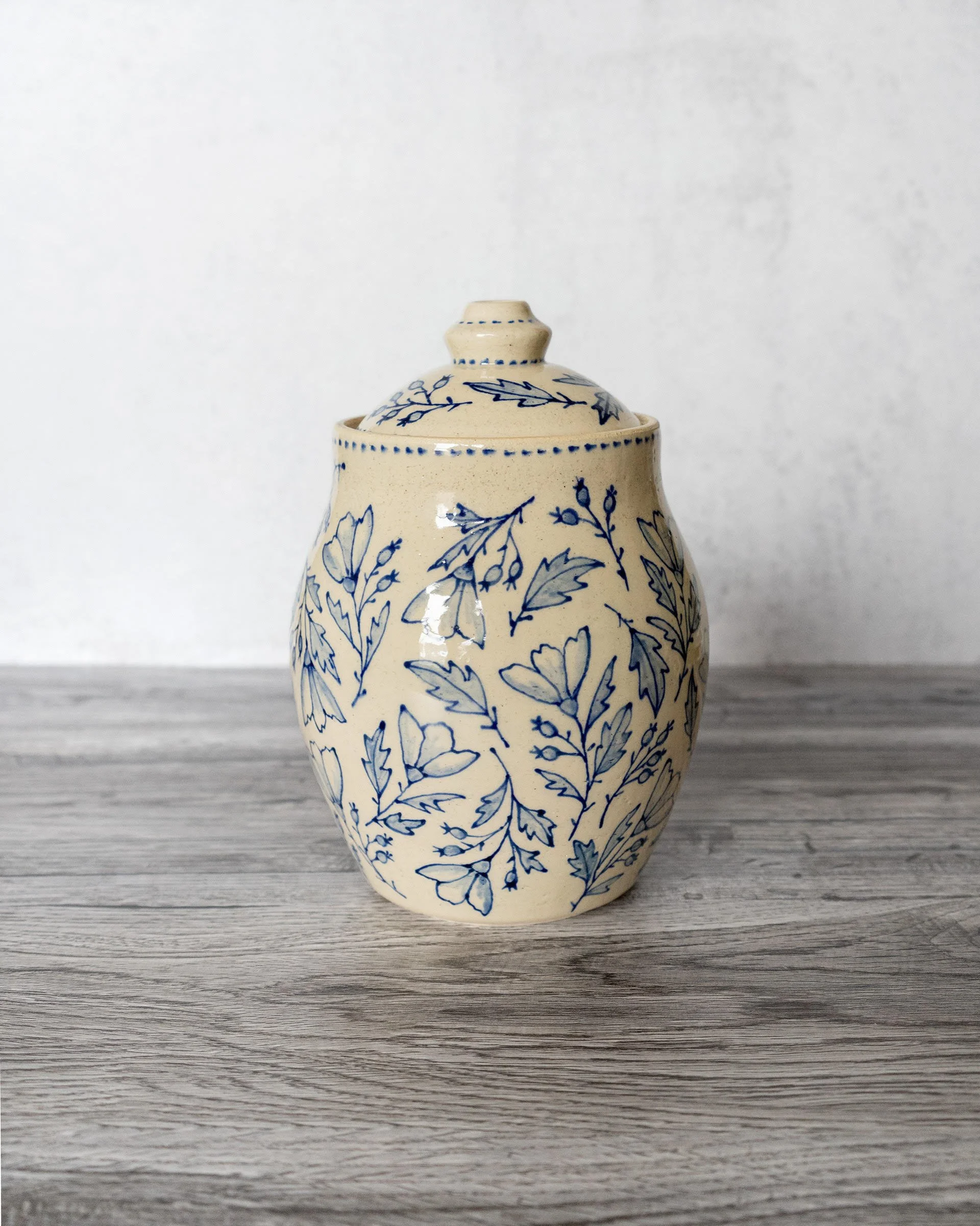 Cream-colored ceramic vase with blue floral pattern, placed on a wooden surface with a plain white background by Amy Brockman