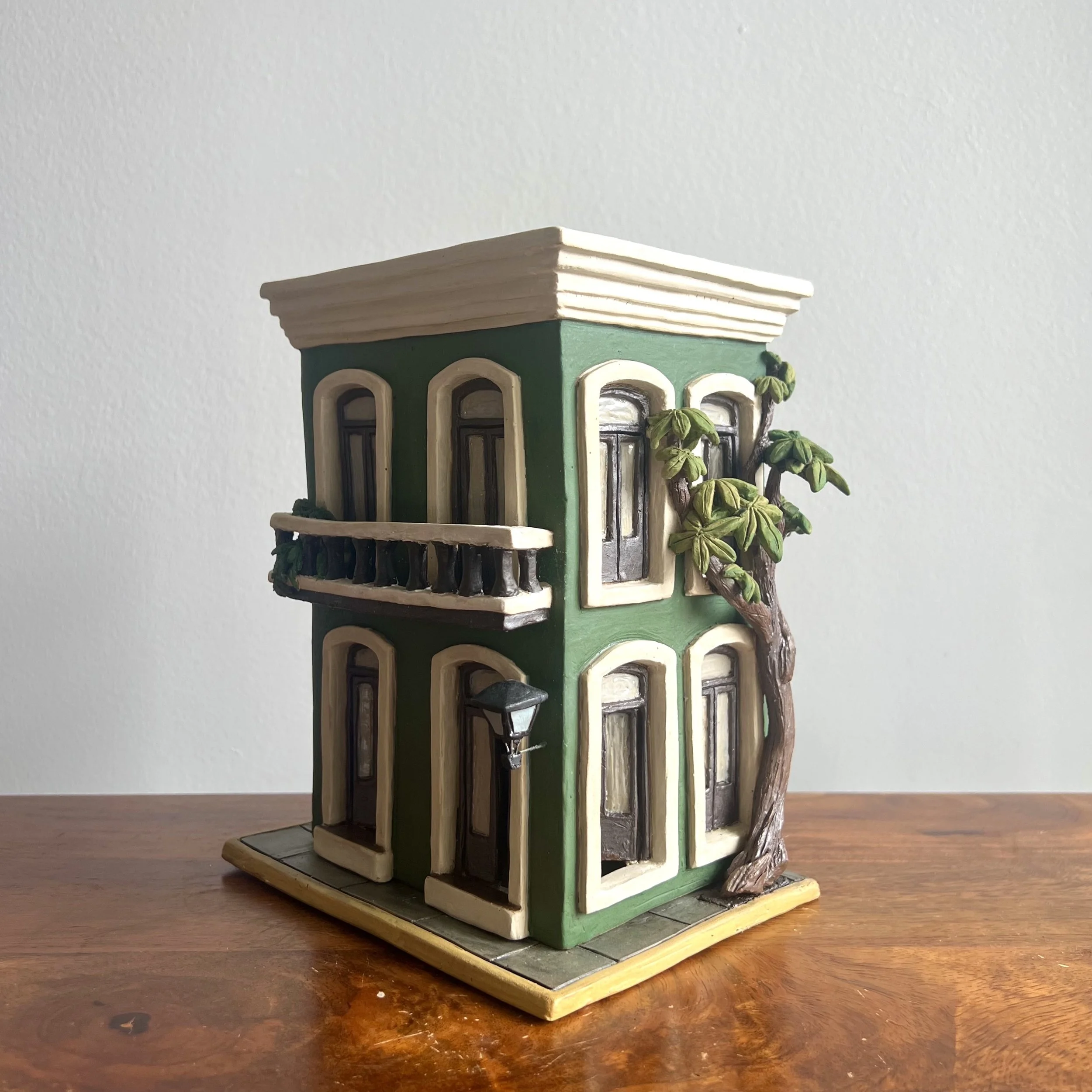 Miniature model of a green, three-story building with beige trim, arched windows, a small balcony with plants, a street lamp, and a tree, placed on a wooden surface against a plain white wall. - Gleidymar Rivera