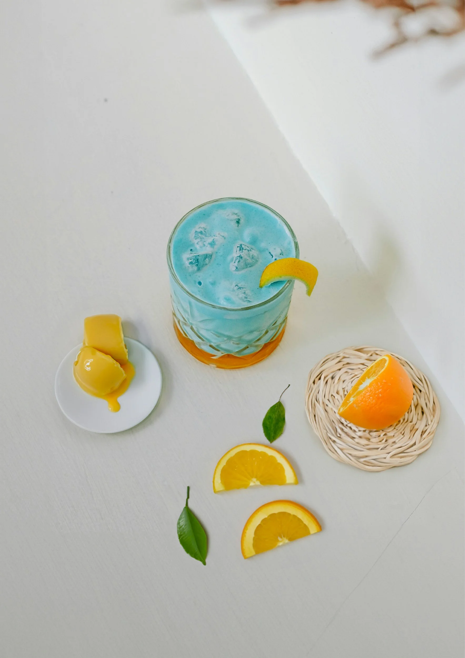 A glass of blue cocktail with ice and a lemon wedge on the rim, surrounded by slices of orange, lemon, green leaves, and a small white plate with lemon slices, all on a white surface.