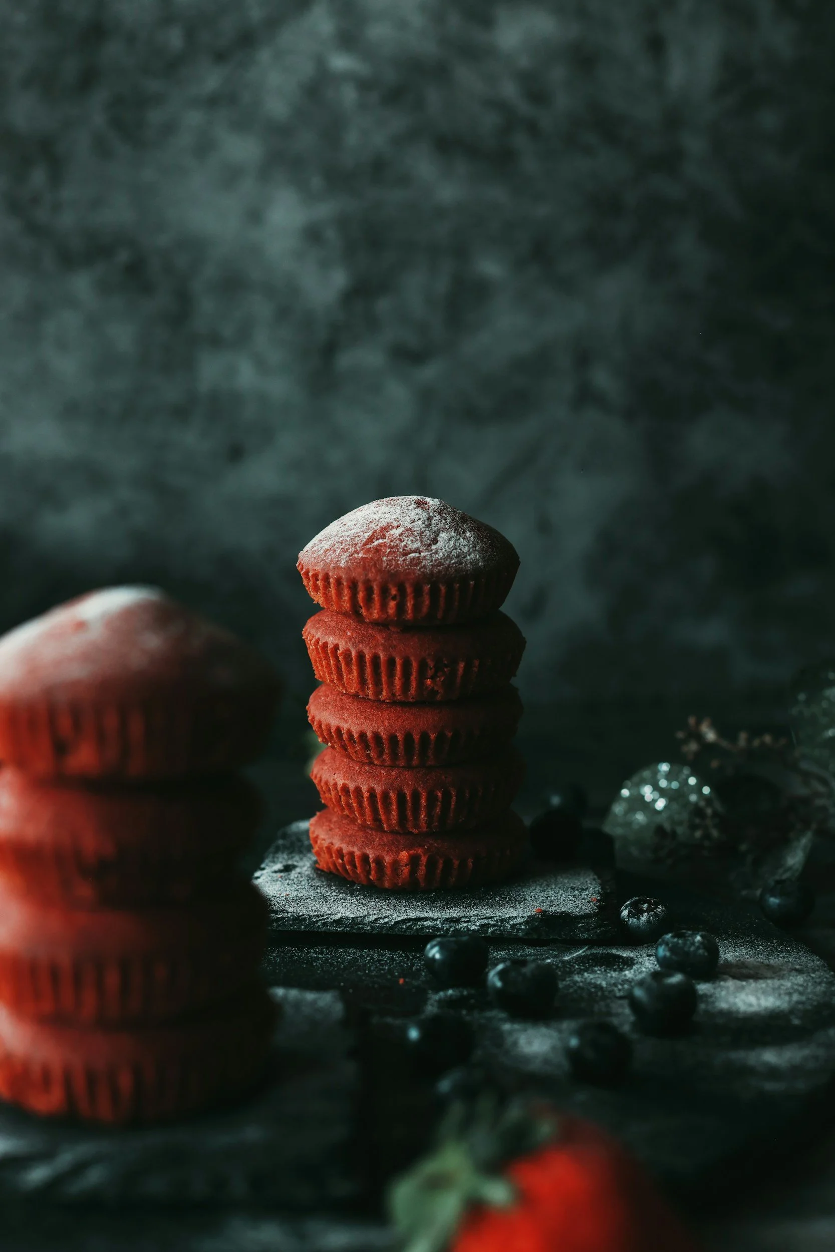 A stack of red cupcakes topped with powdered sugar, with a blurred foreground of additional cupcakes and berries, against a dark textured background.
