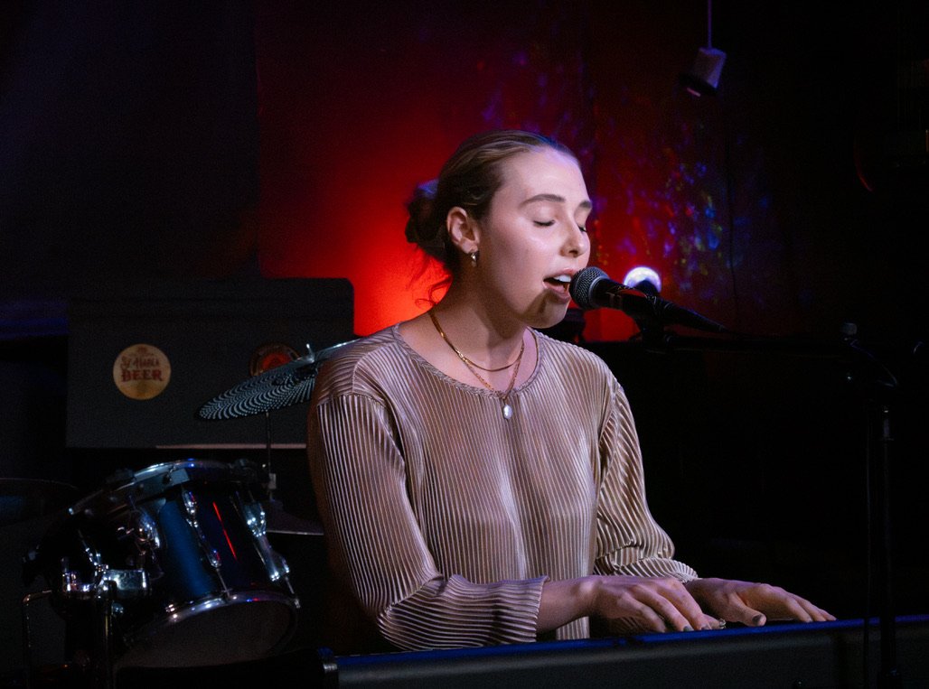 Independent singer-songwriter Jennifer B. Porter performing at the piano in a live music venue with red and purple stage lighting.