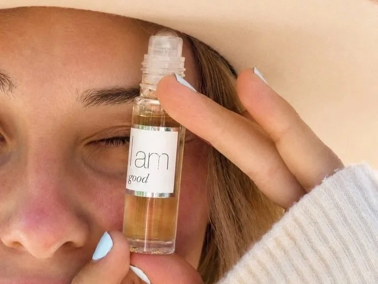I AM Fragrance Good perfume bottle held beside a woman's face in a close-up lifestyle campaign image.